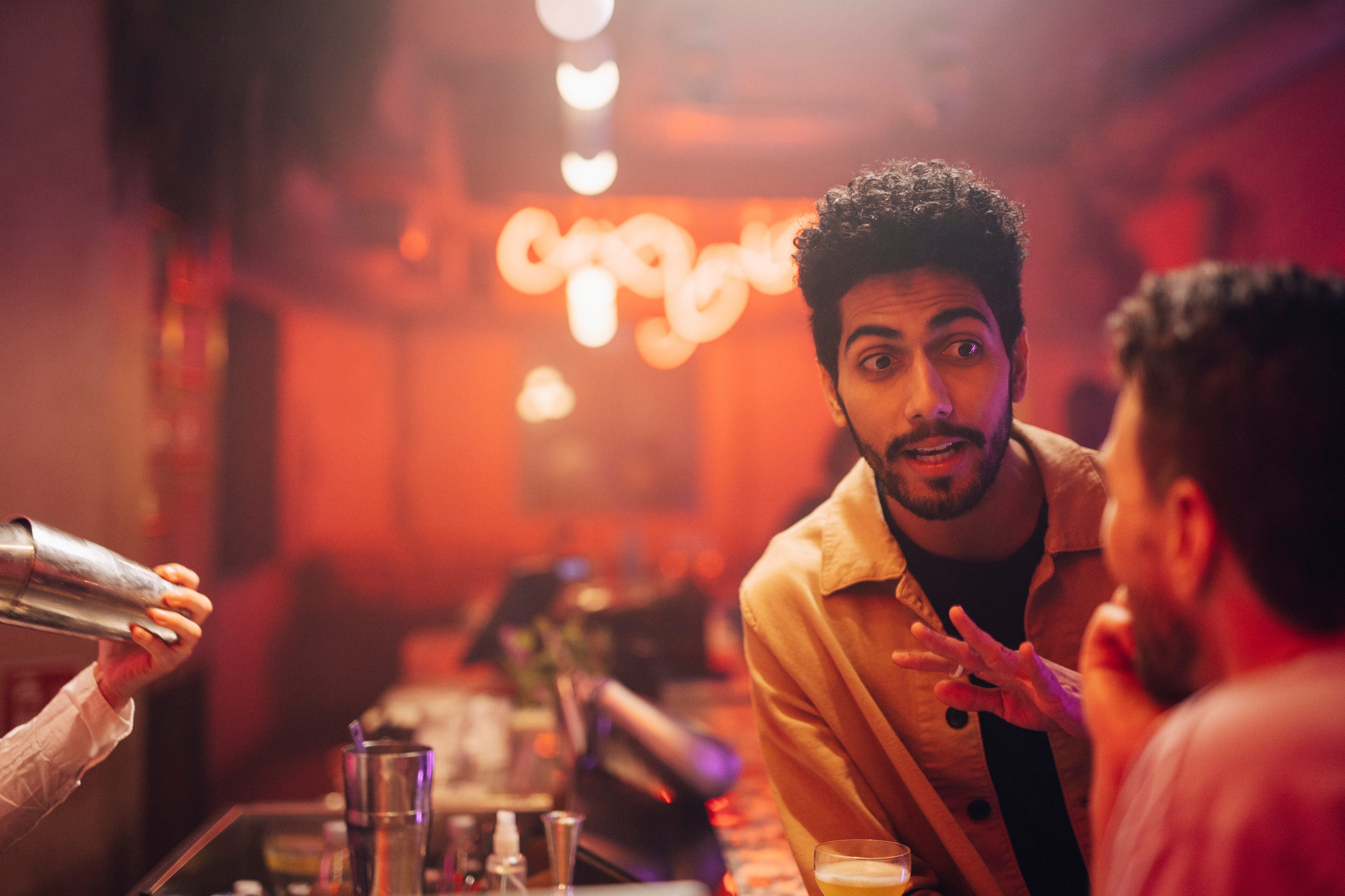 Tre personer chatter på en svakt opplyst bar, en gestikulerer animert. En bartender tilbereder en drink i nærheten