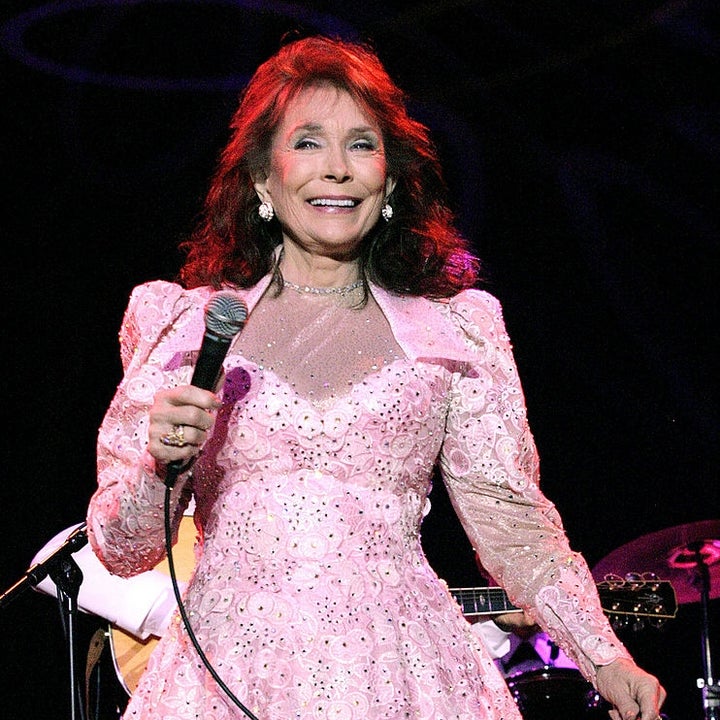 A person is on stage, smiling and holding a microphone, wearing an intricate, long-sleeved, lace dress