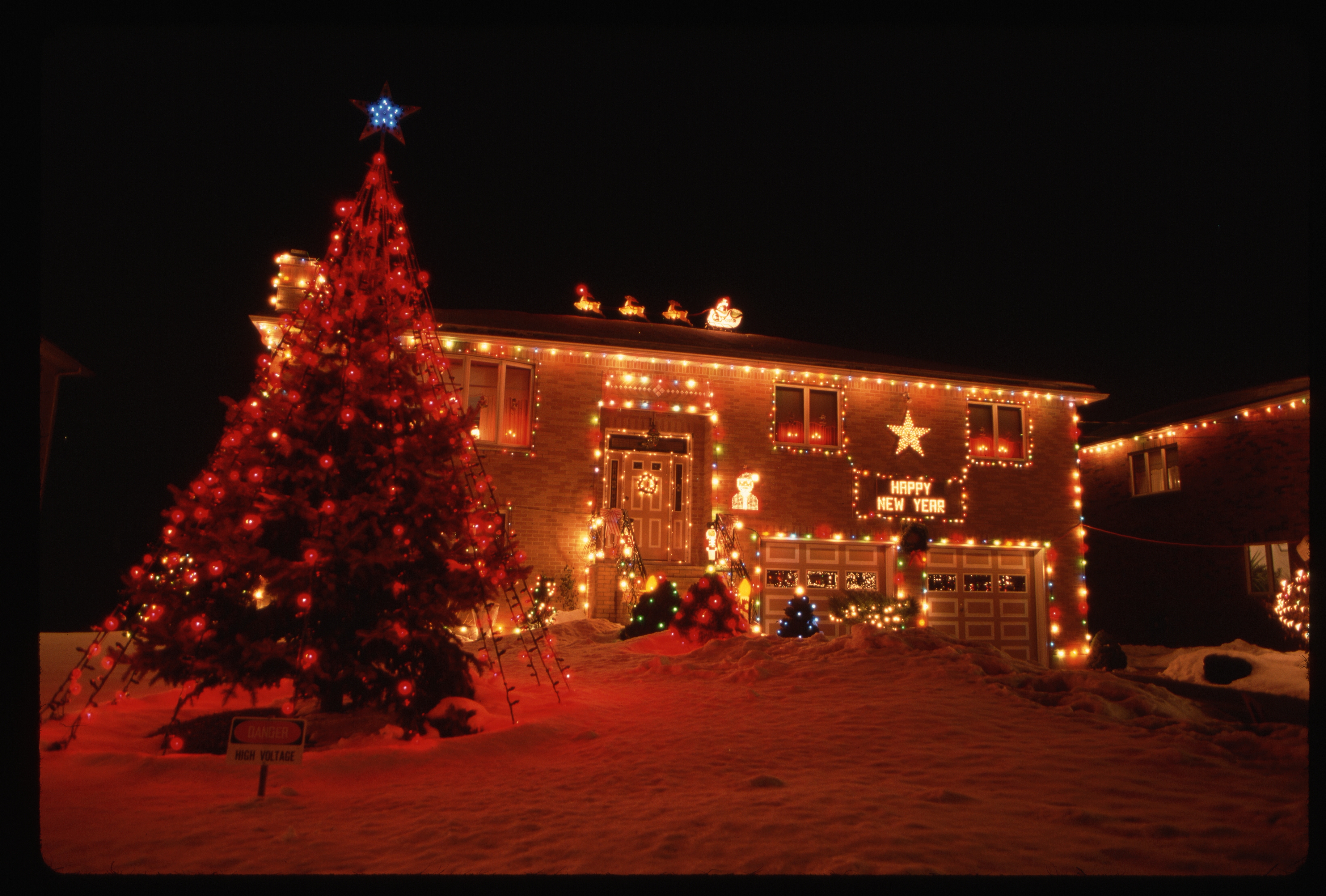 House with festive holiday lights, large decorated tree outside, and a "Happy New Year" sign on the front. Snow covers the ground