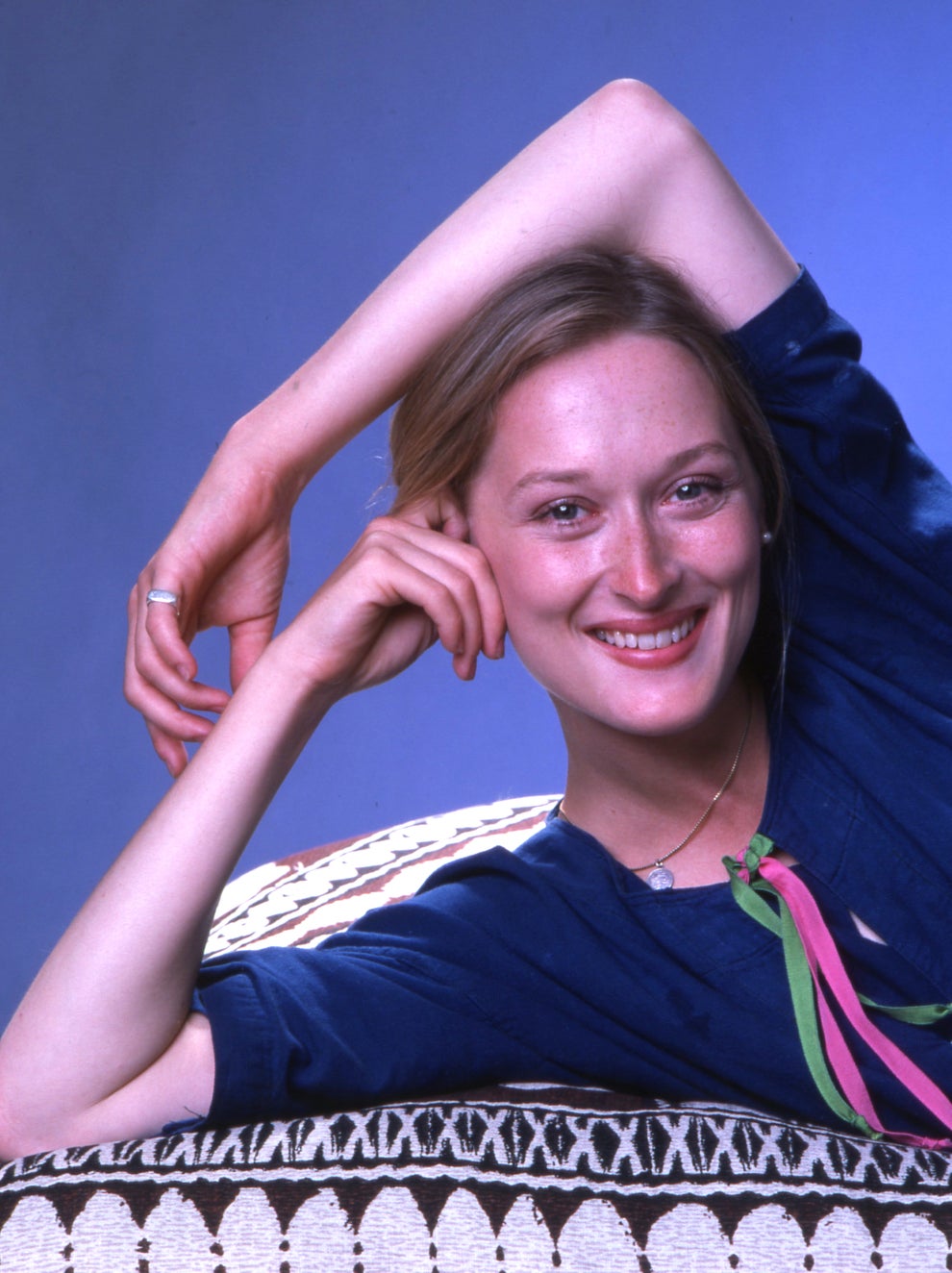 21 Jaw-Dropping "Then And Now" Photos Of Celebrities Who Have Gotten Wayyyyy Sexier With Age 4 Person lounging on a patterned couch, smiling with a relaxed hand above head. Wearing casual clothing with a colorful ribbon detail