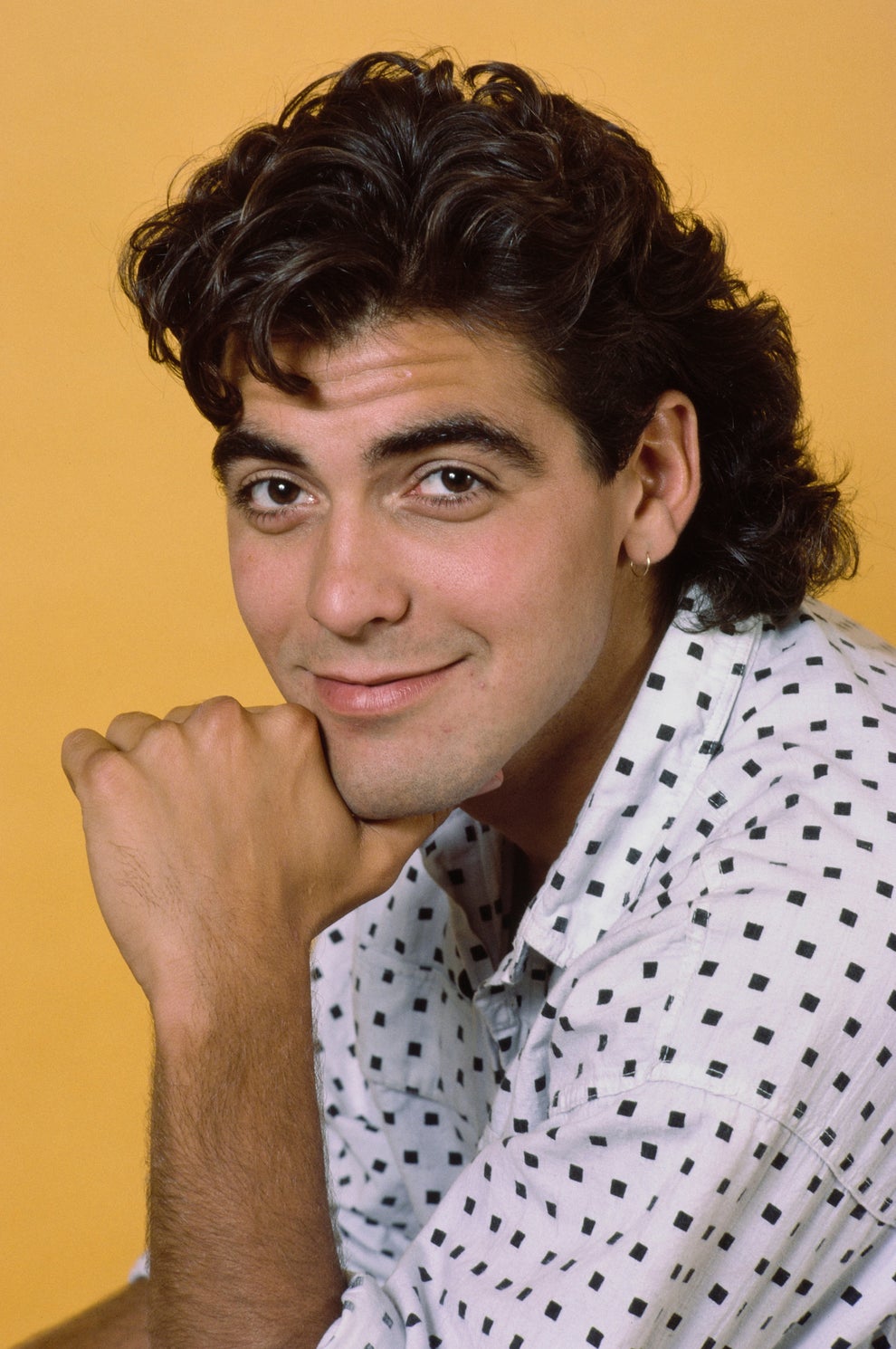21 Jaw-Dropping "Then And Now" Photos Of Celebrities Who Have Gotten Wayyyyy Sexier With Age 24 Person with a mullet hairstyle, wearing a patterned shirt, smiles with chin resting on hand against a plain background