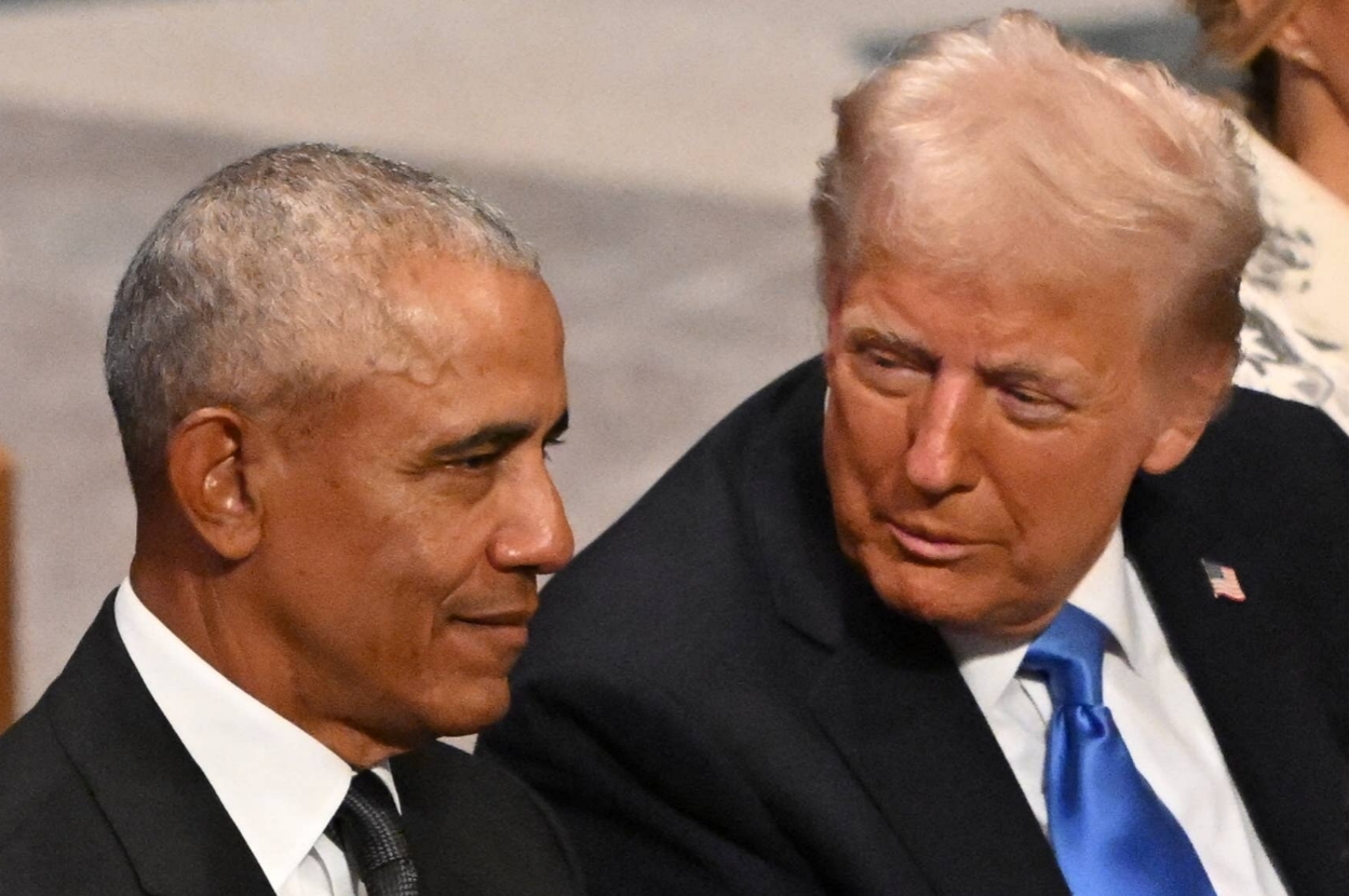 Two men in suits sit closely, engaged in conversation at an event, with one appearing to listen attentively while the other speaks