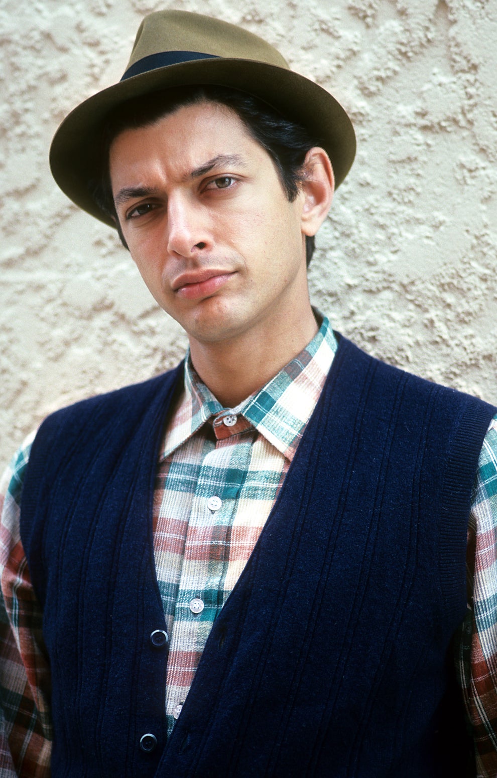21 Jaw-Dropping "Then And Now" Photos Of Celebrities Who Have Gotten Wayyyyy Sexier With Age 70 Person wearing a plaid shirt, dark vest, and fedora, posing against a textured wall