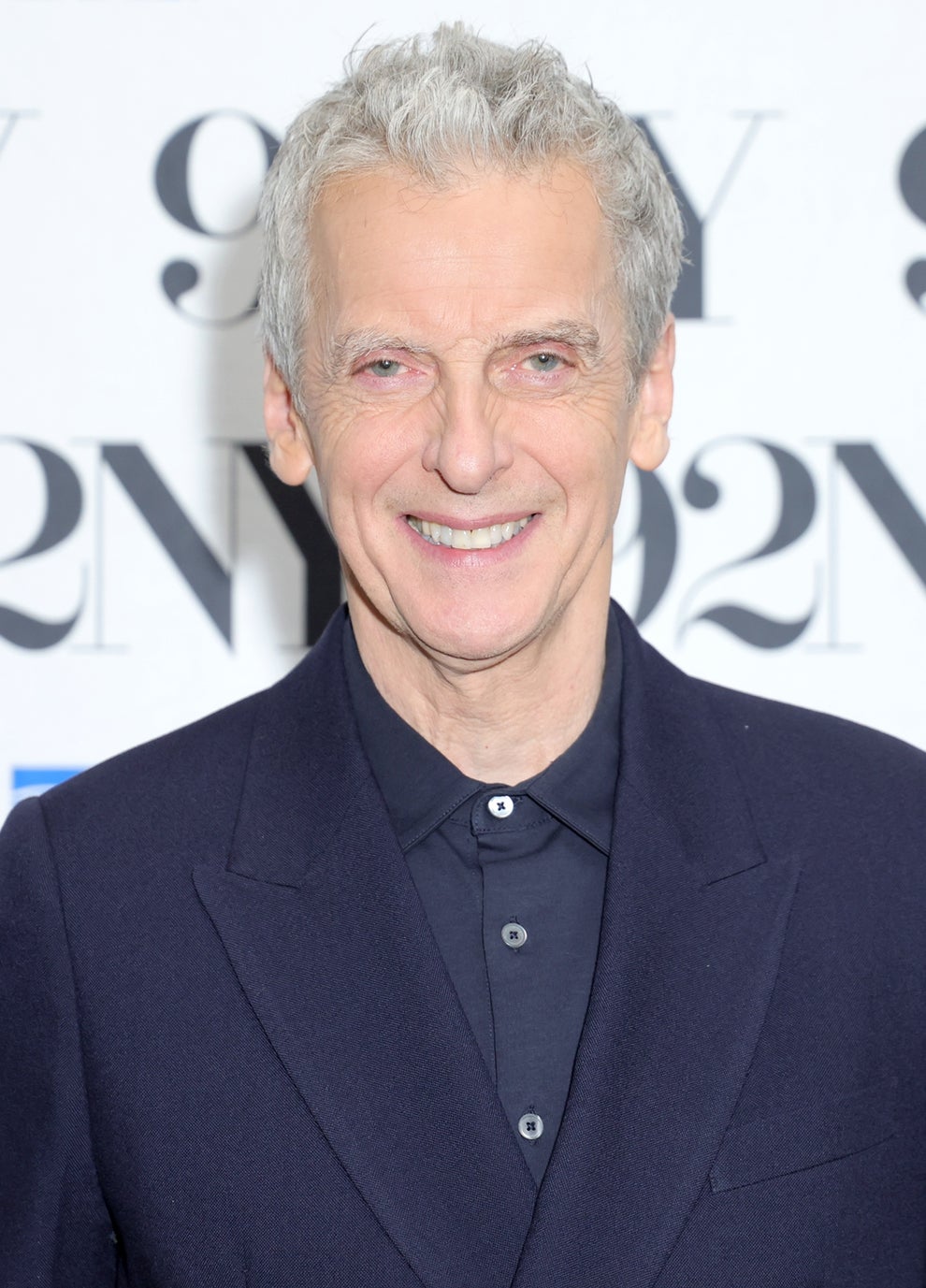 21 Jaw-Dropping "Then And Now" Photos Of Celebrities Who Have Gotten Wayyyyy Sexier With Age 80 Person smiling at an event, wearing a dark suit and shirt, standing in front of a backdrop with "92NY" and sponsor logos