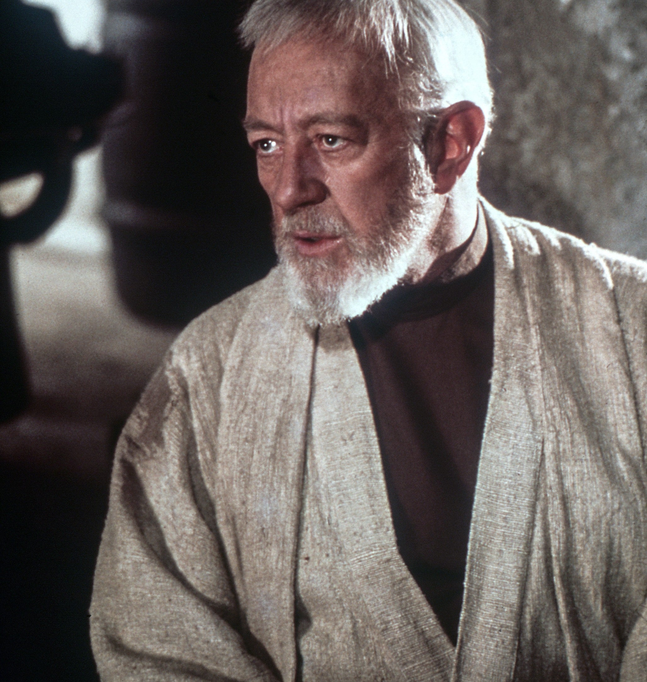 An older man with a white beard and robe appears thoughtful, sitting indoors