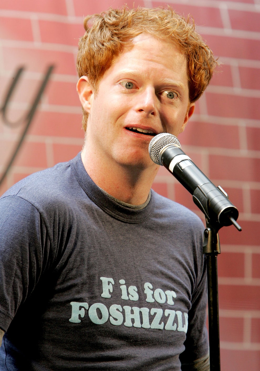 21 Jaw-Dropping "Then And Now" Photos Of Celebrities Who Have Gotten Wayyyyy Sexier With Age 74 Person with short hair in a casual T-shirt, reading "F is for FOSHIZZLE," speaking into a microphone on stage