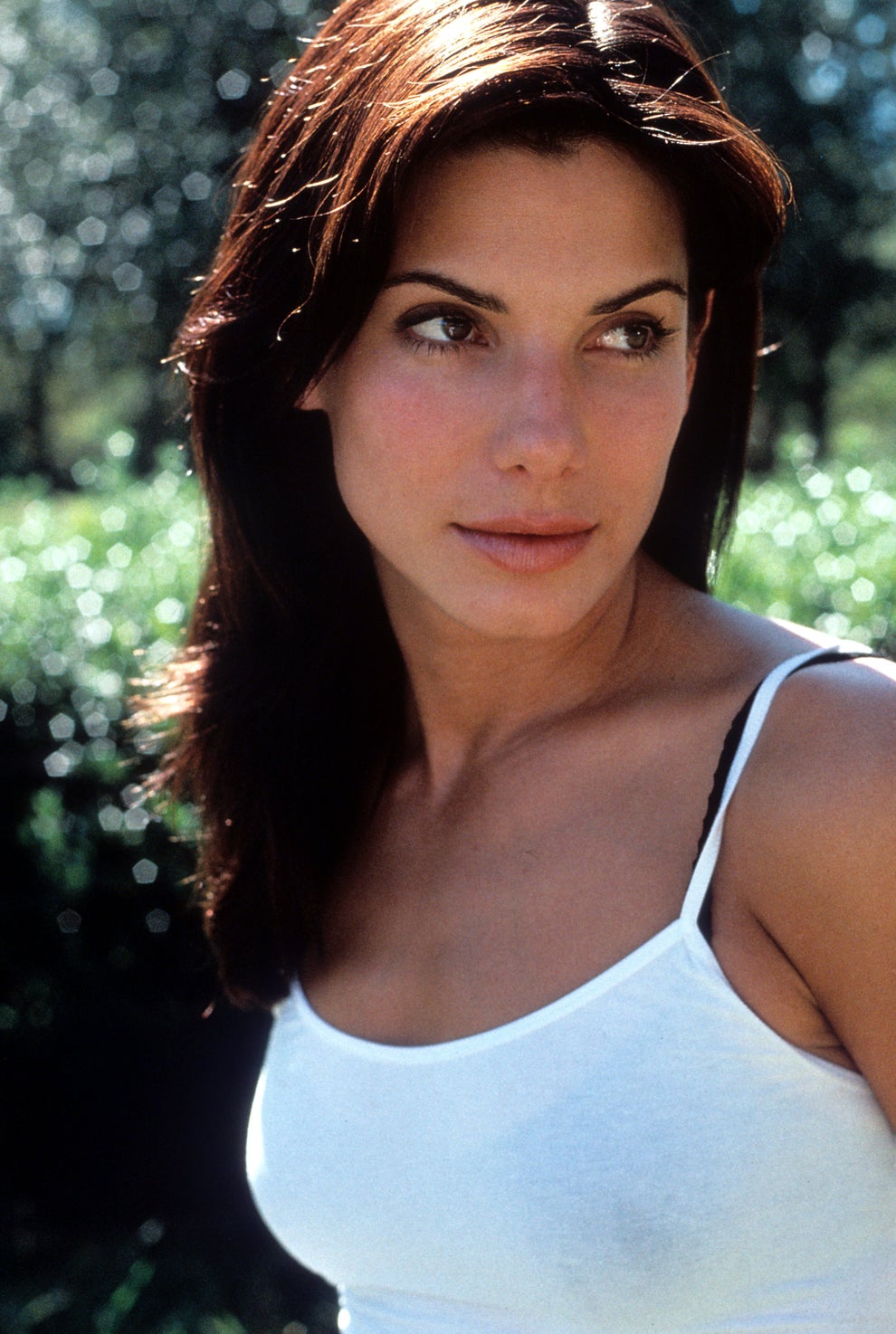 21 Jaw-Dropping "Then And Now" Photos Of Celebrities Who Have Gotten Wayyyyy Sexier With Age 66 Person outdoors wearing a white tank top, looking to the side with a neutral expression. Trees are blurred in the background