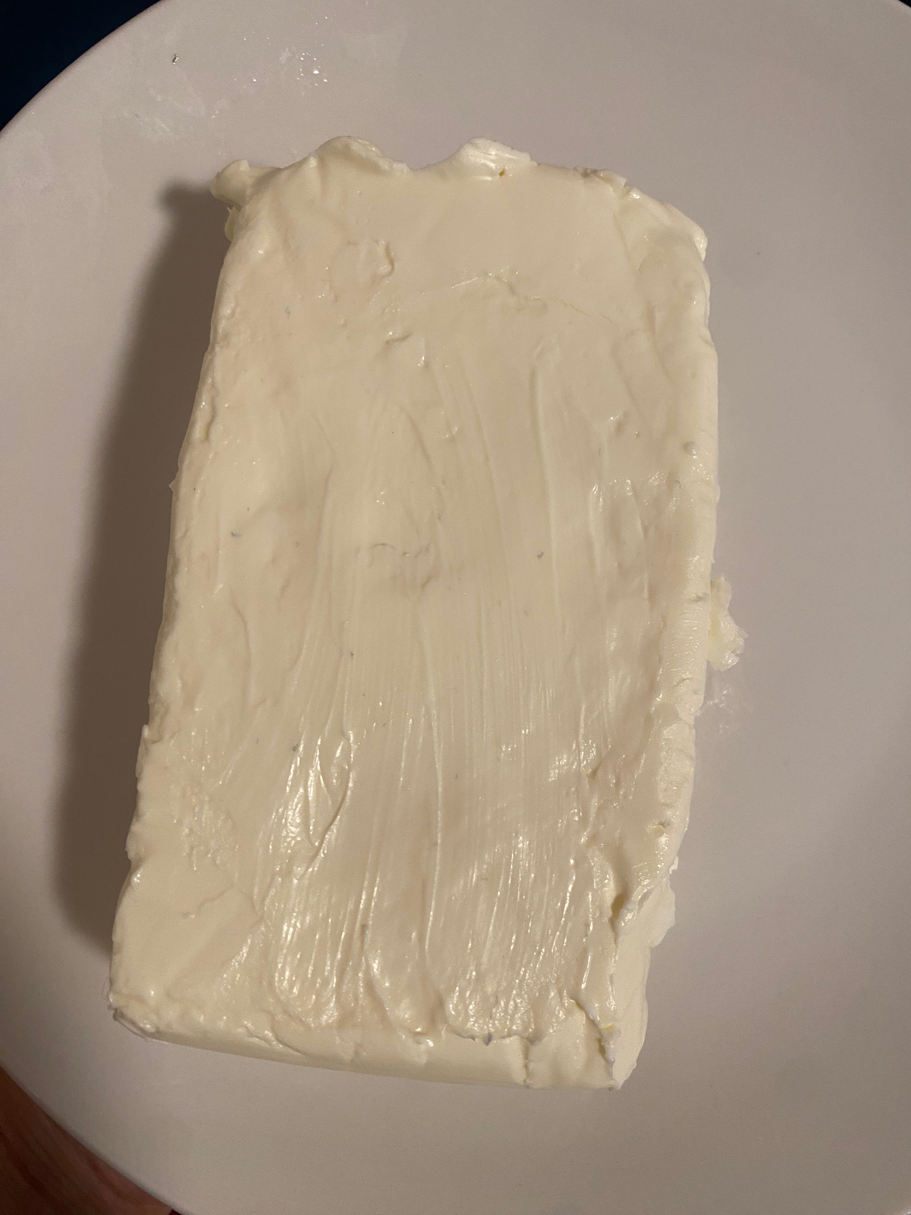 A rectangular block of cream cheese placed on a round white plate. The surface appears smooth and creamy