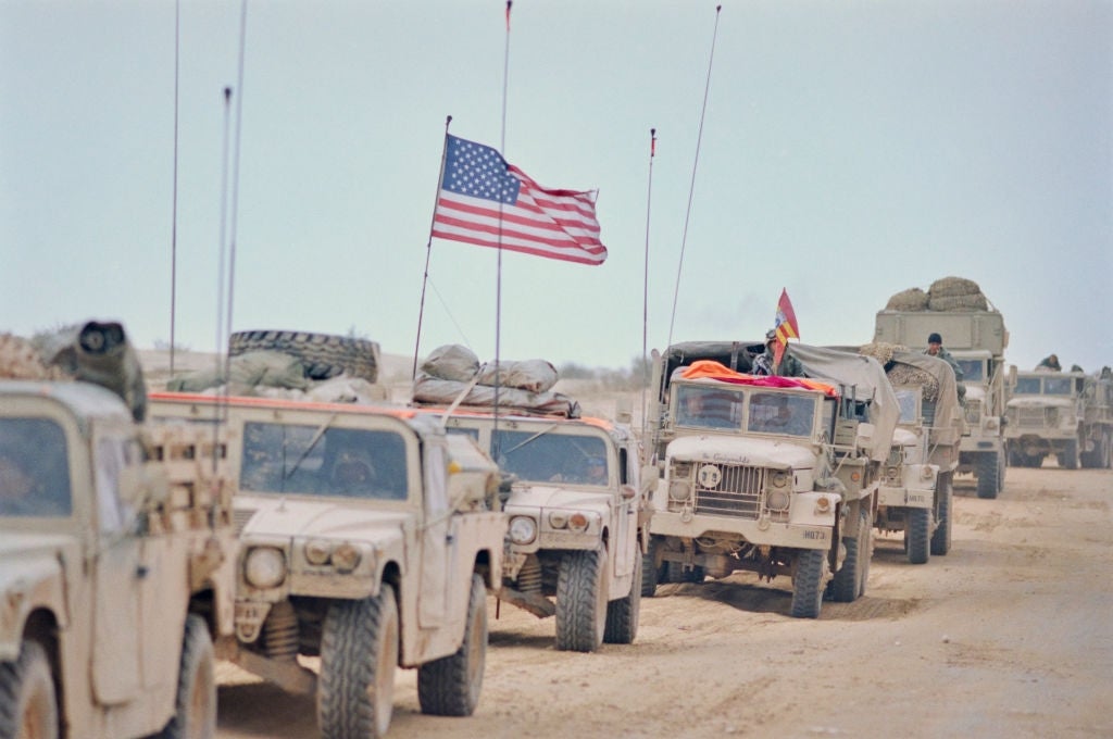 Military convoy of transport vehicles with American flag, traveling done  a godforsaken  landscape. Clusters of cogwheel  are secured connected  the vehicles