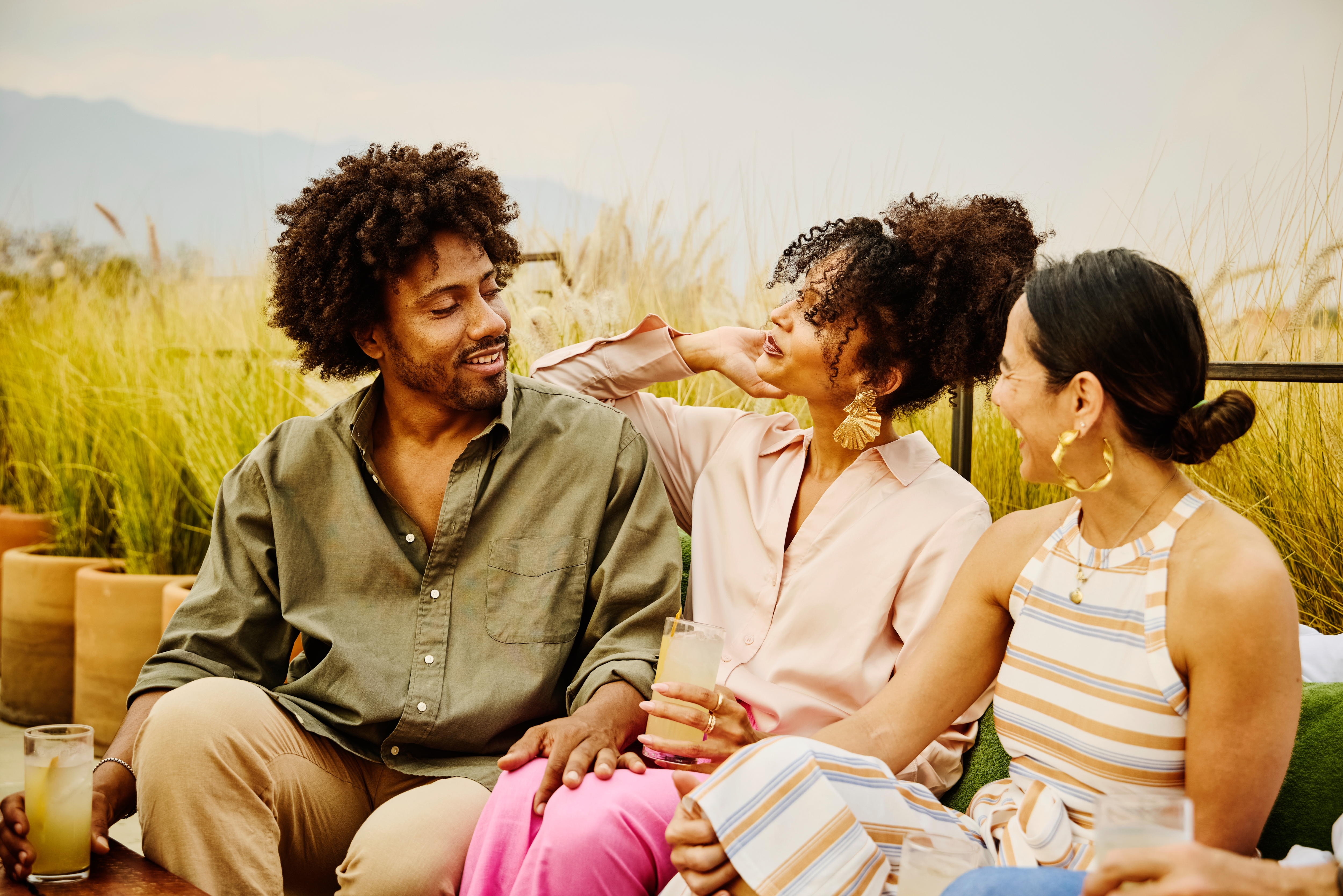 Three people sit outside, smiling and enjoying drinks together, suggesting a relaxed, friendly atmosphere