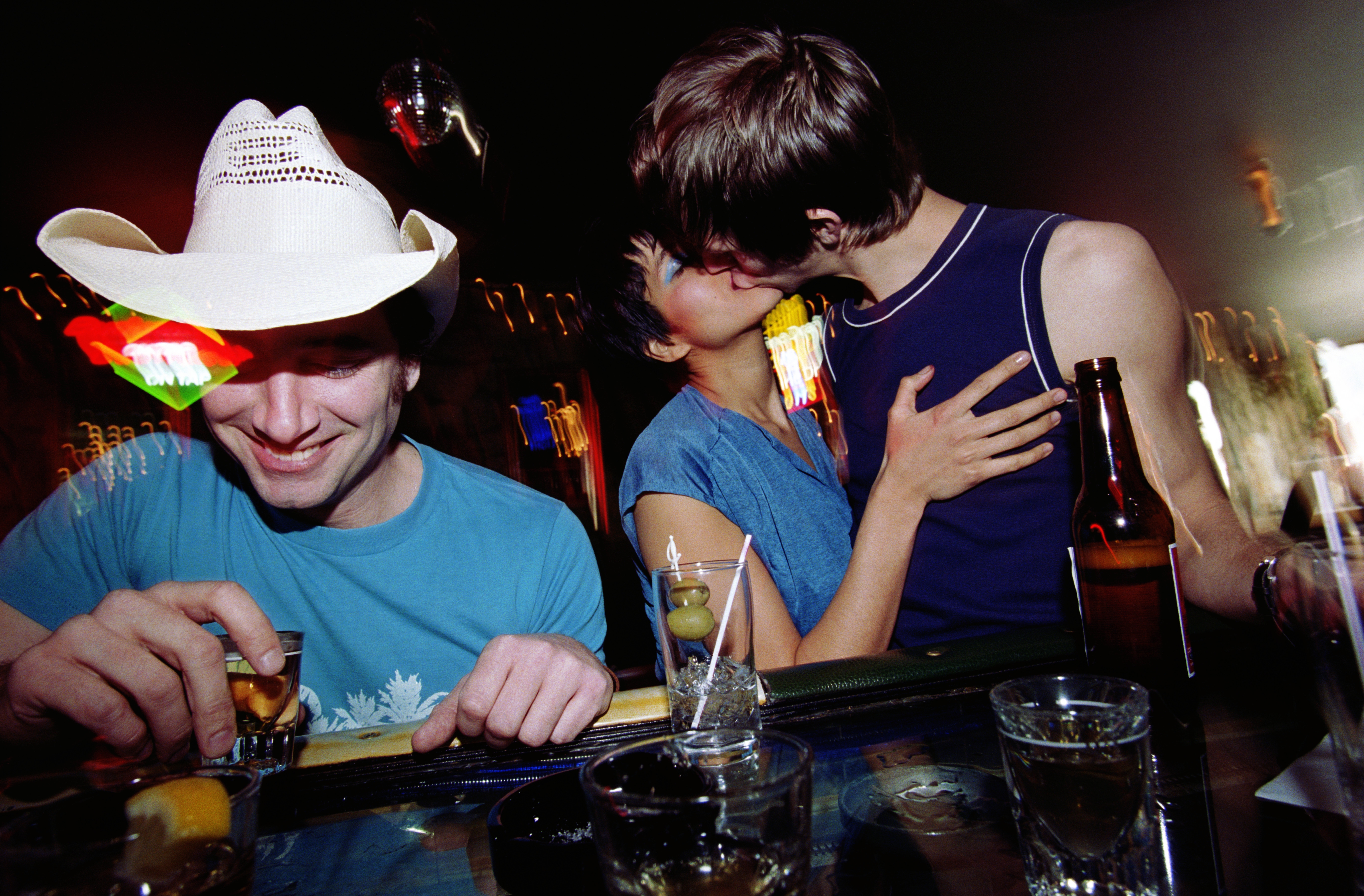 A couple kisses passionately at a bar, while a person in a cowboy hat laughs with a drink
