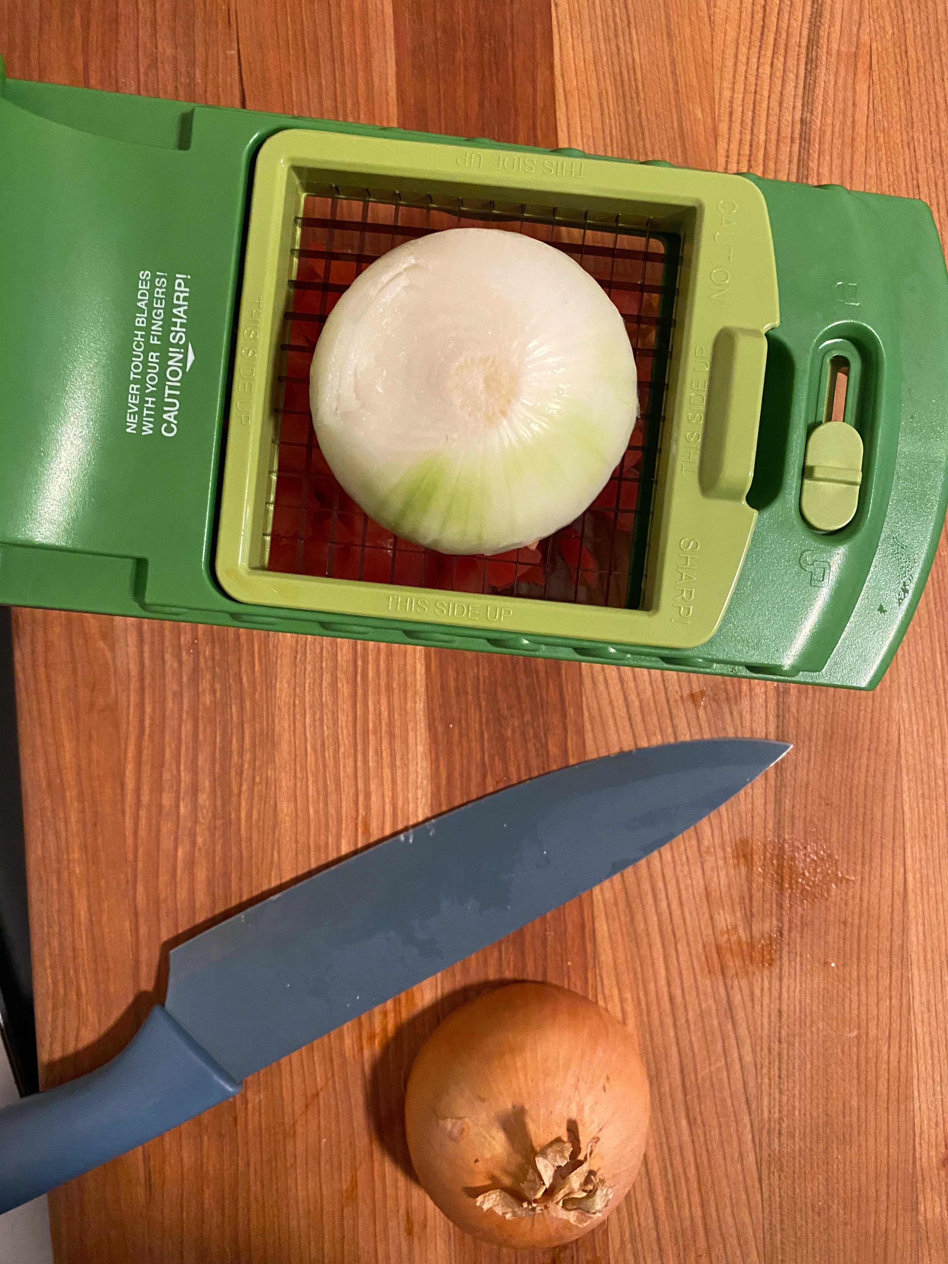 Kitchen tool with an onion being sliced placed on a cutting board, adjacent to a knife and an unsliced onion