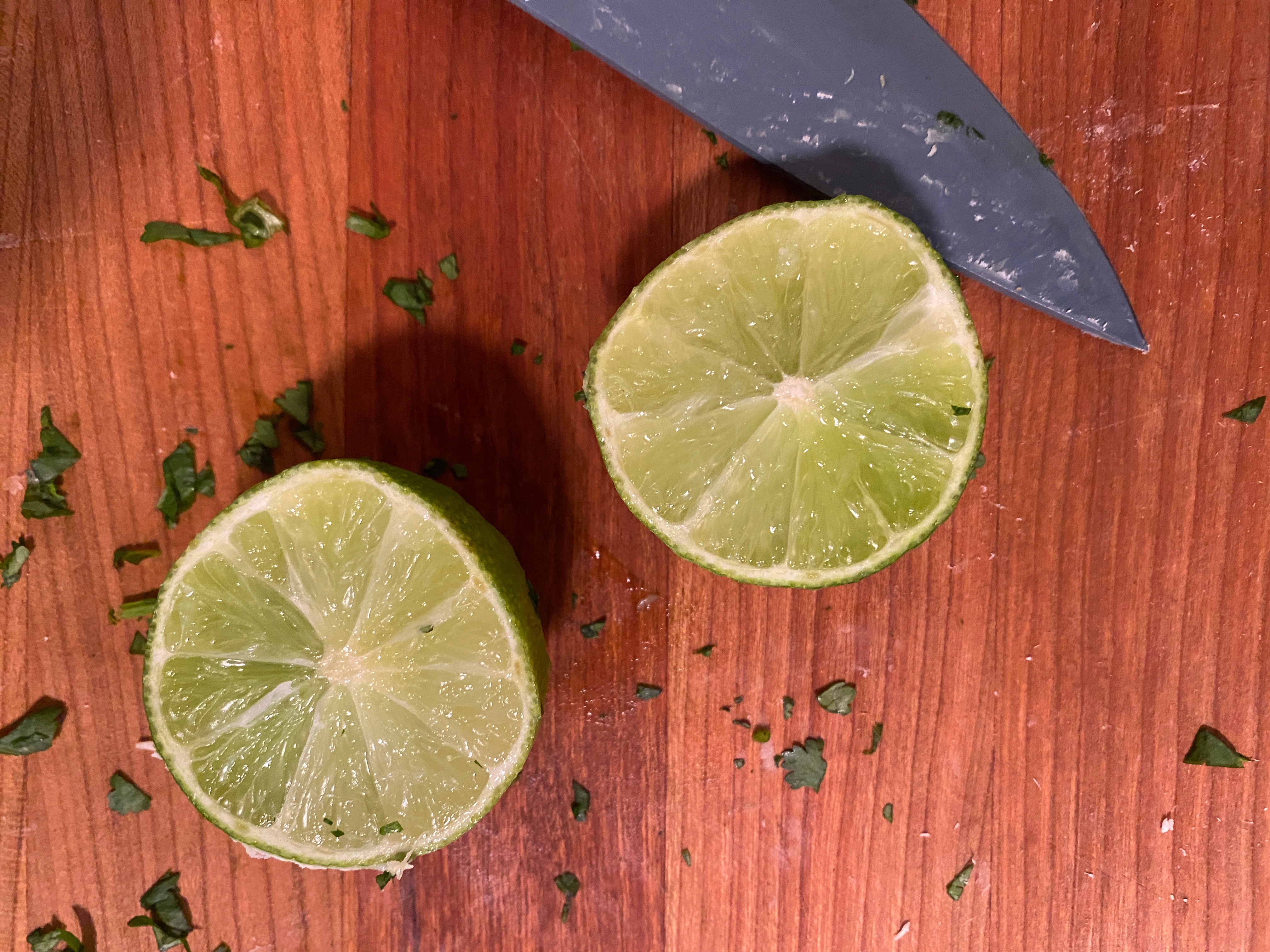 Two lime halves on a wooden surface with a knife and scattered herb pieces nearby