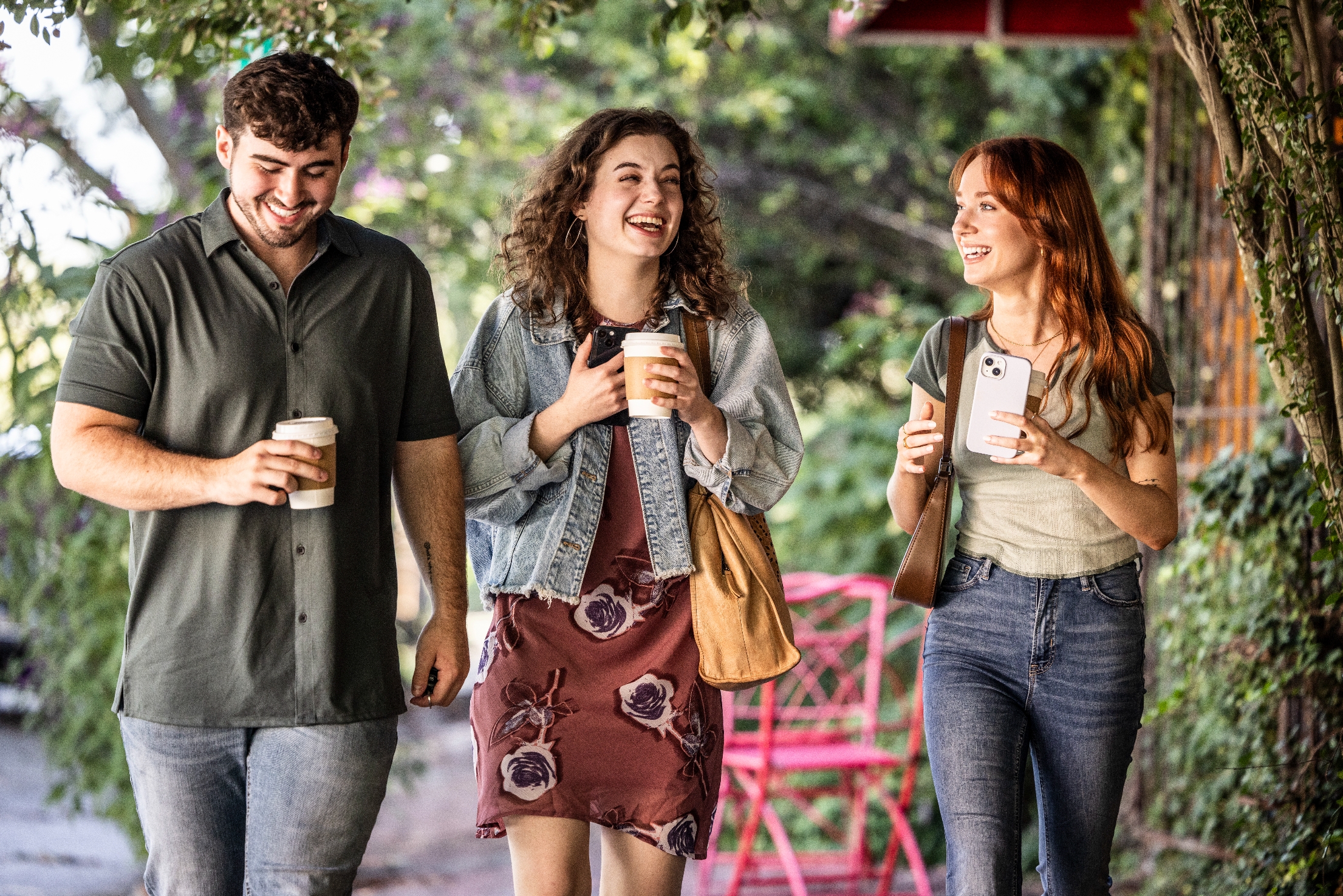 Three friends walk with coffee, smiling and chatting outdoors. One wears a denim jacket, another a polo shirt, and the third has a casual top and jeans