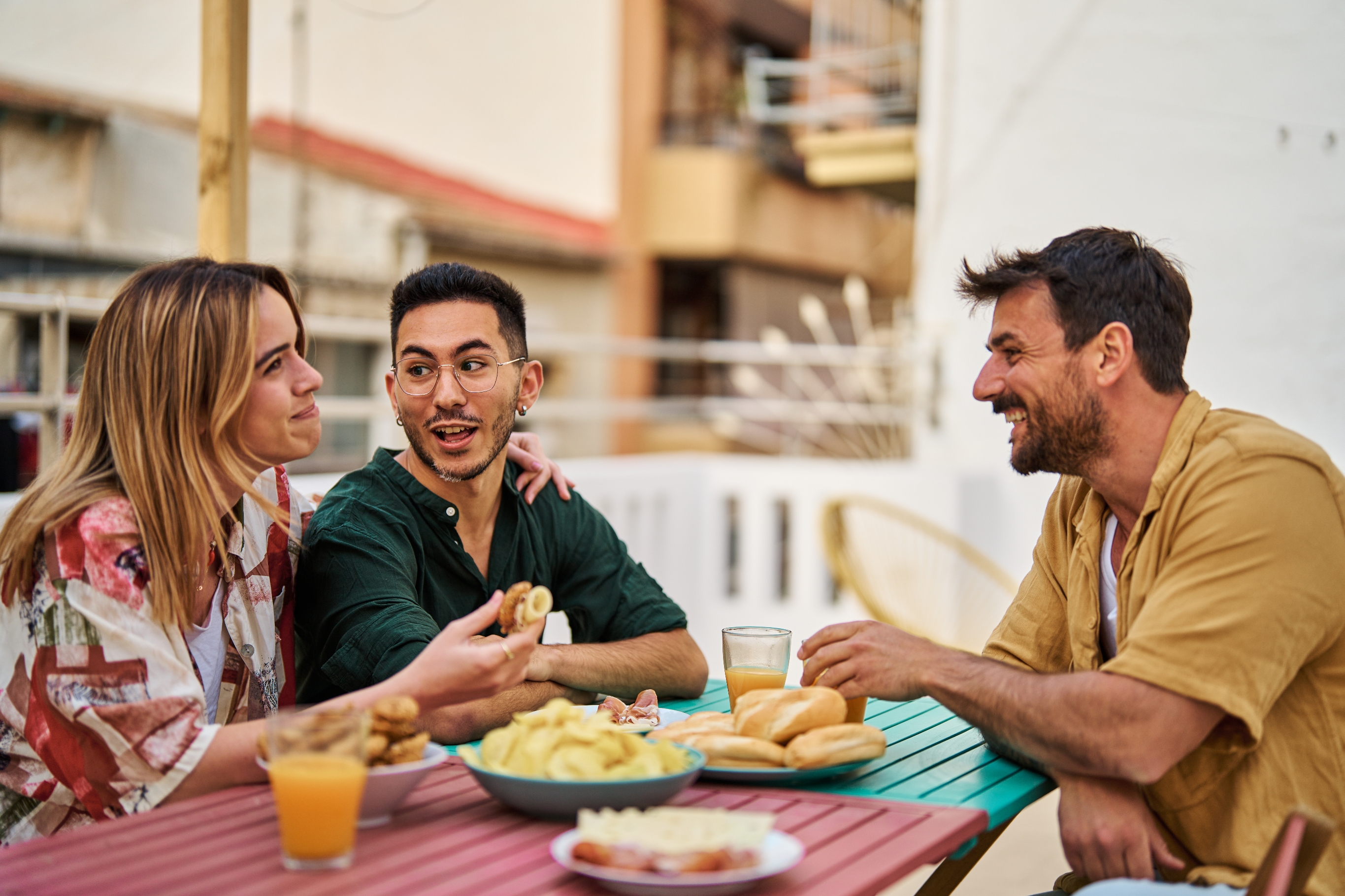Three people sitting at a table outdoors, smiling and chatting over snacks and drinks, suggesting a casual, friendly gathering