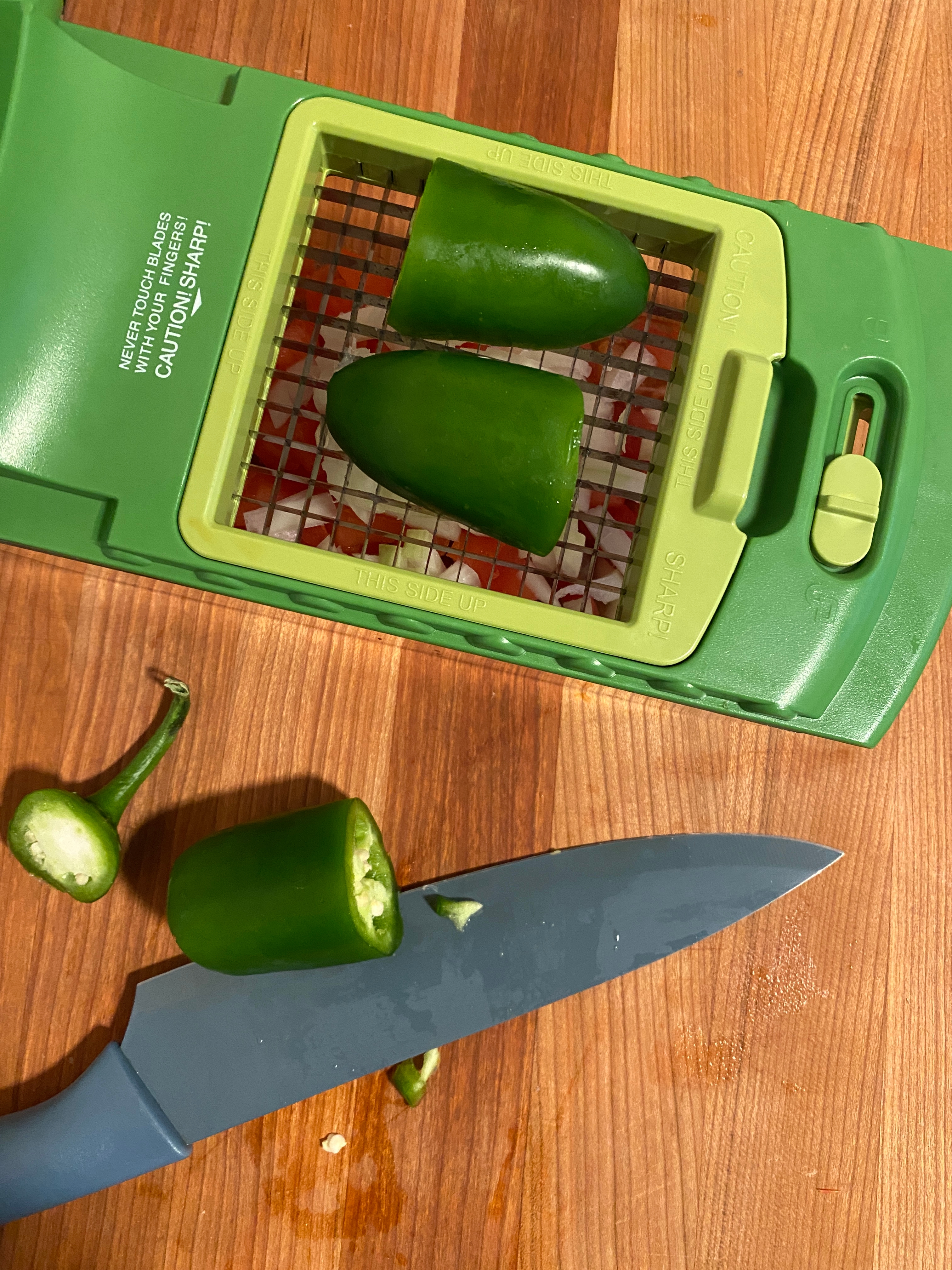 Jalapeño halves on a vegetable slicer with a blue knife nearby on a wooden surface. Whole pepper and stem on side
