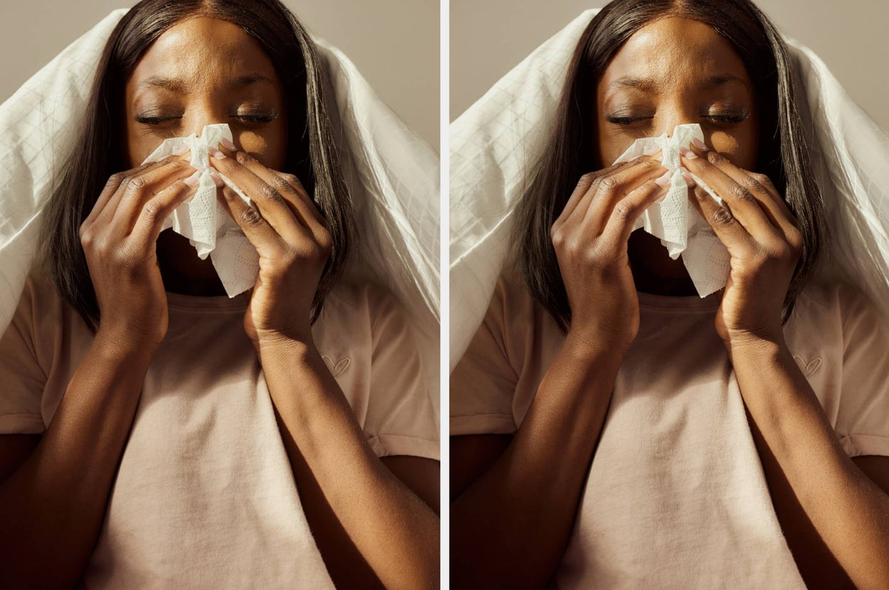 Person in a casual t-shirt sneezing into a tissue, eyes closed, with a blanket over their head, implying a cold or allergy symptoms