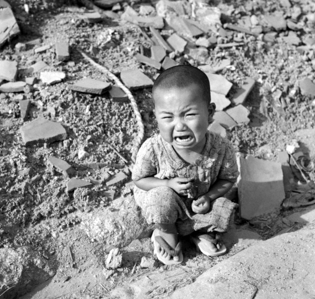 A young kid  sits among rubble, crying, barefoot, aft  the weaponry  dropped