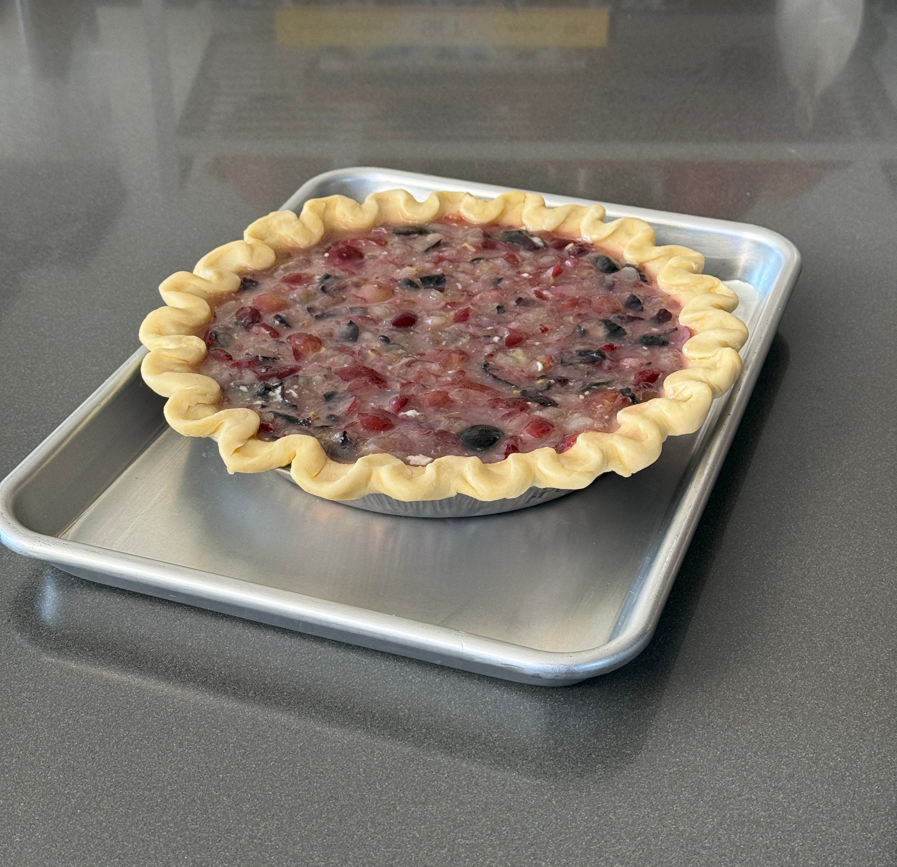 Unbaked grape pie with a crimped-edge crust sits on a baking sheet, ready for the oven