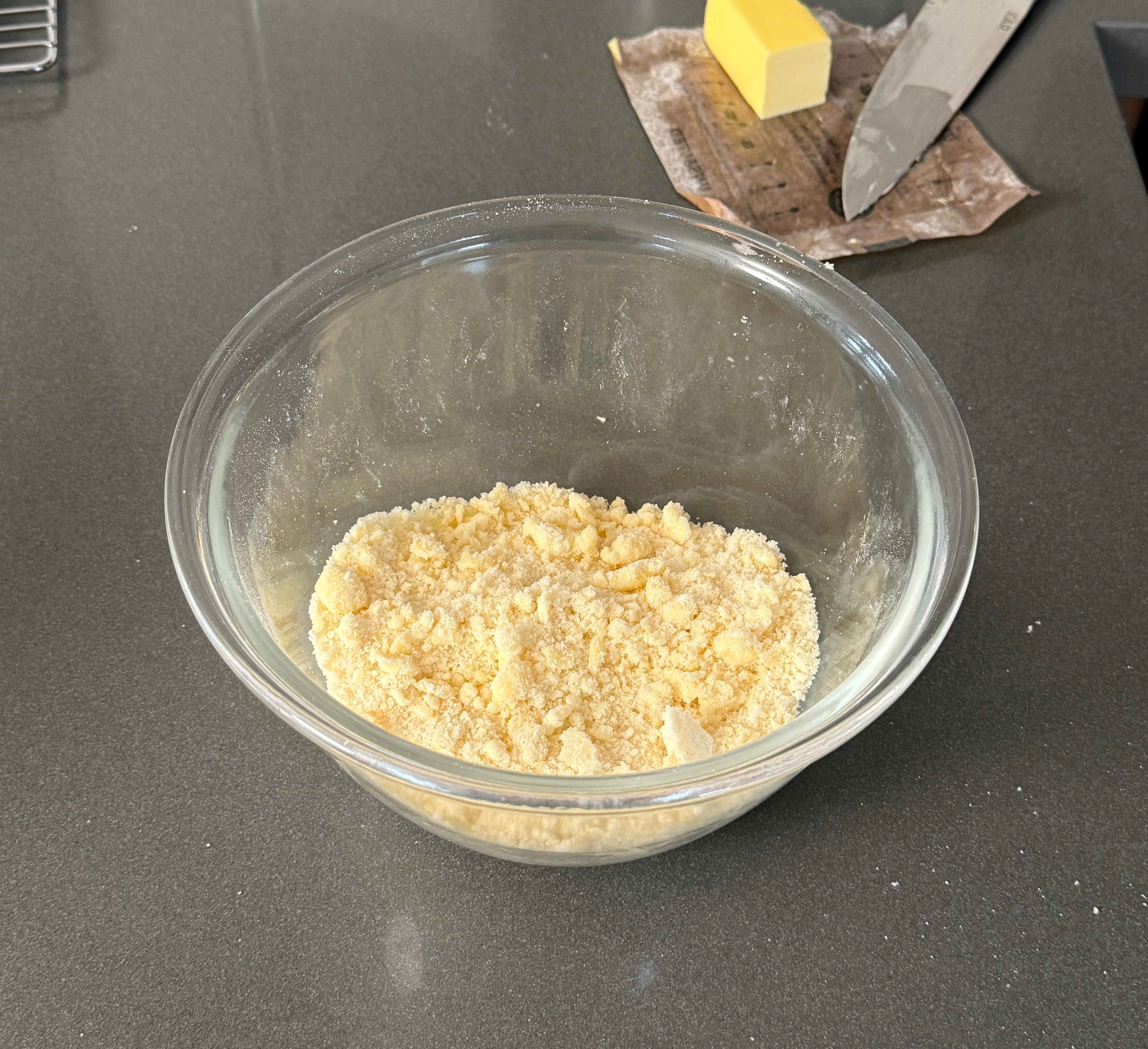 Glass bowl with crumbly mixture on a kitchen counter. Nearby are butter, a knife, and a metal rack