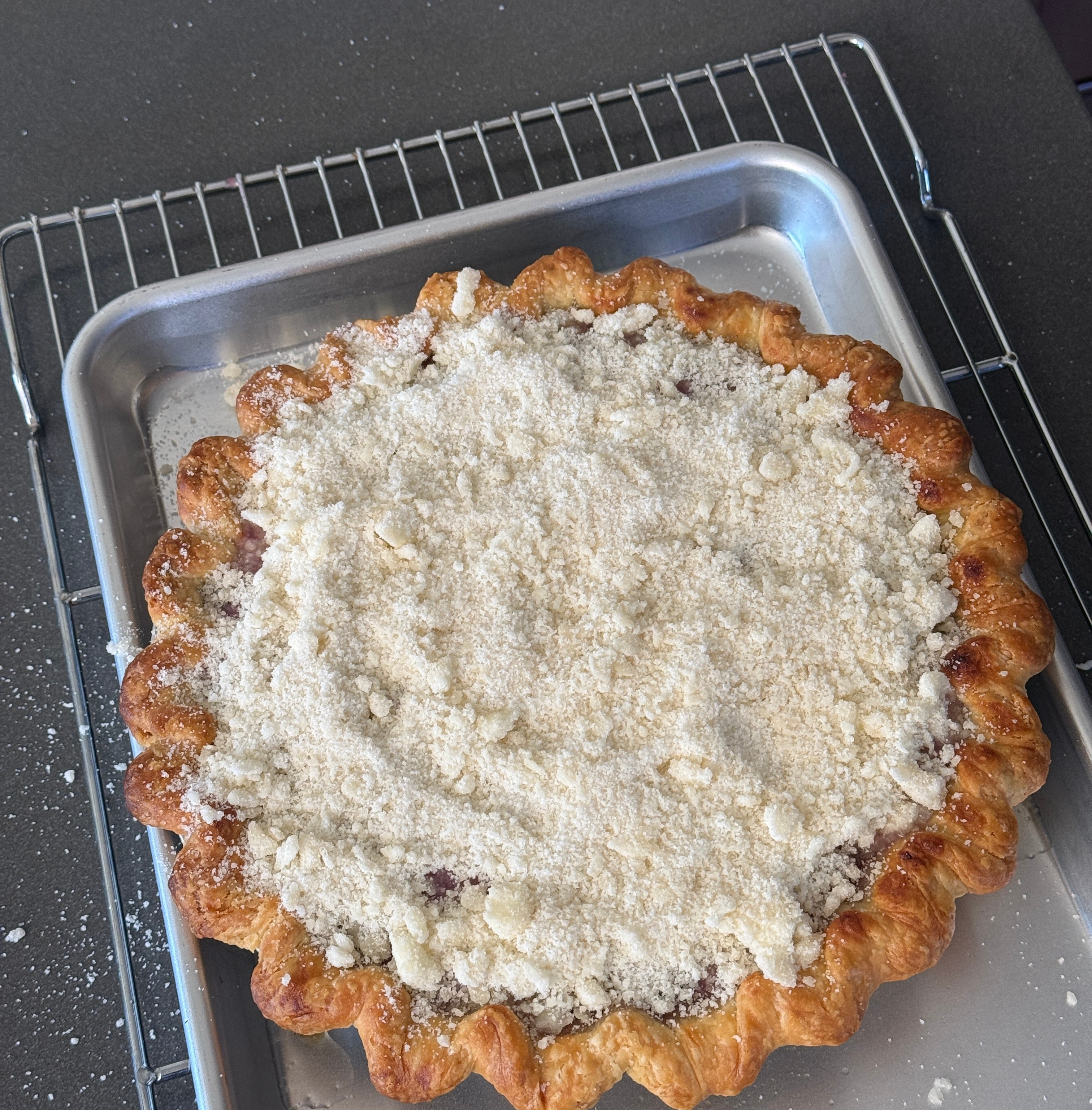 Pie with crumbly topping on a cooling tray set over a baking sheet. Fluted crust edges are visible