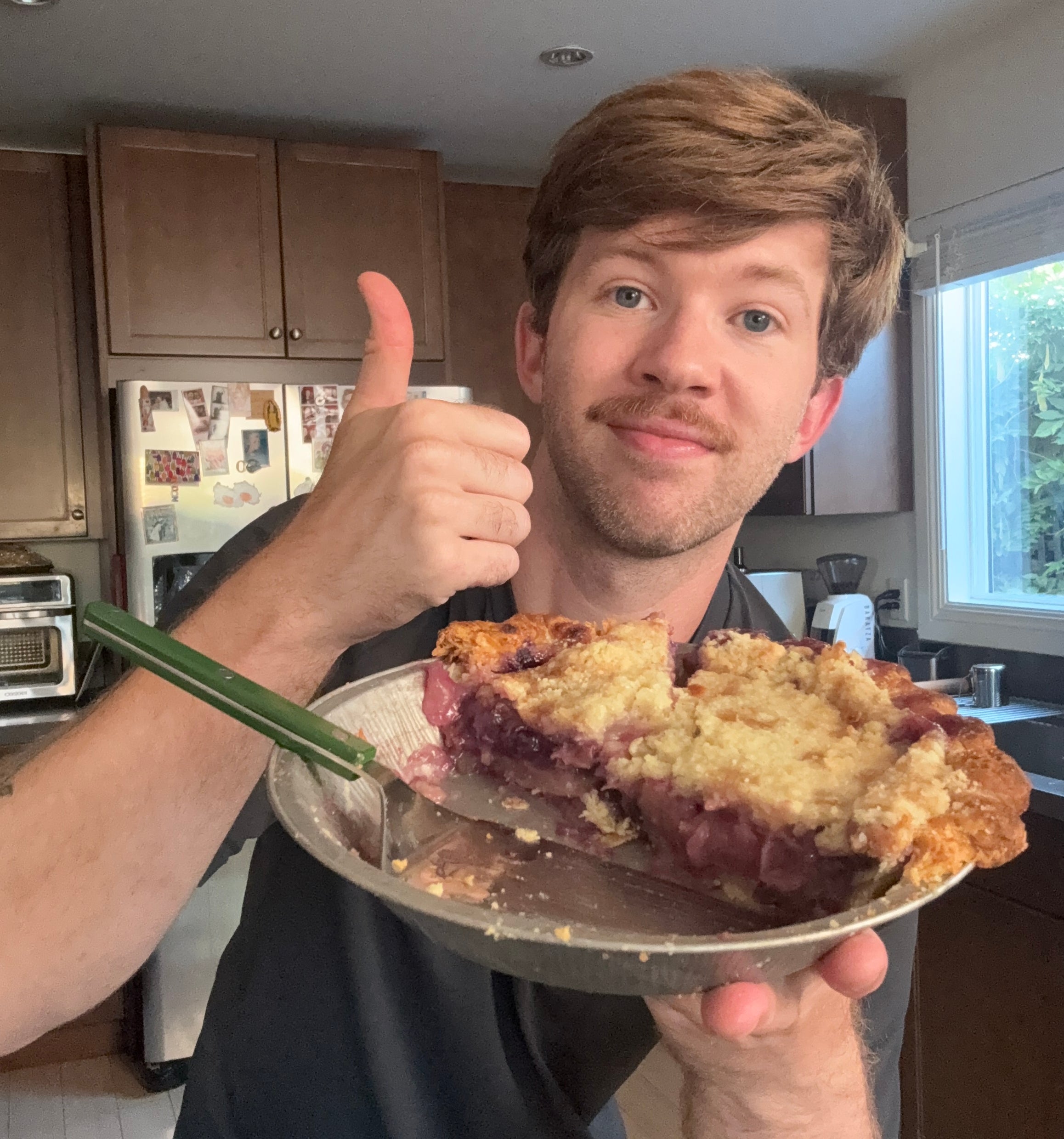 Person in kitchen giving thumbs up while holding a partially eaten pie with a crumb topping