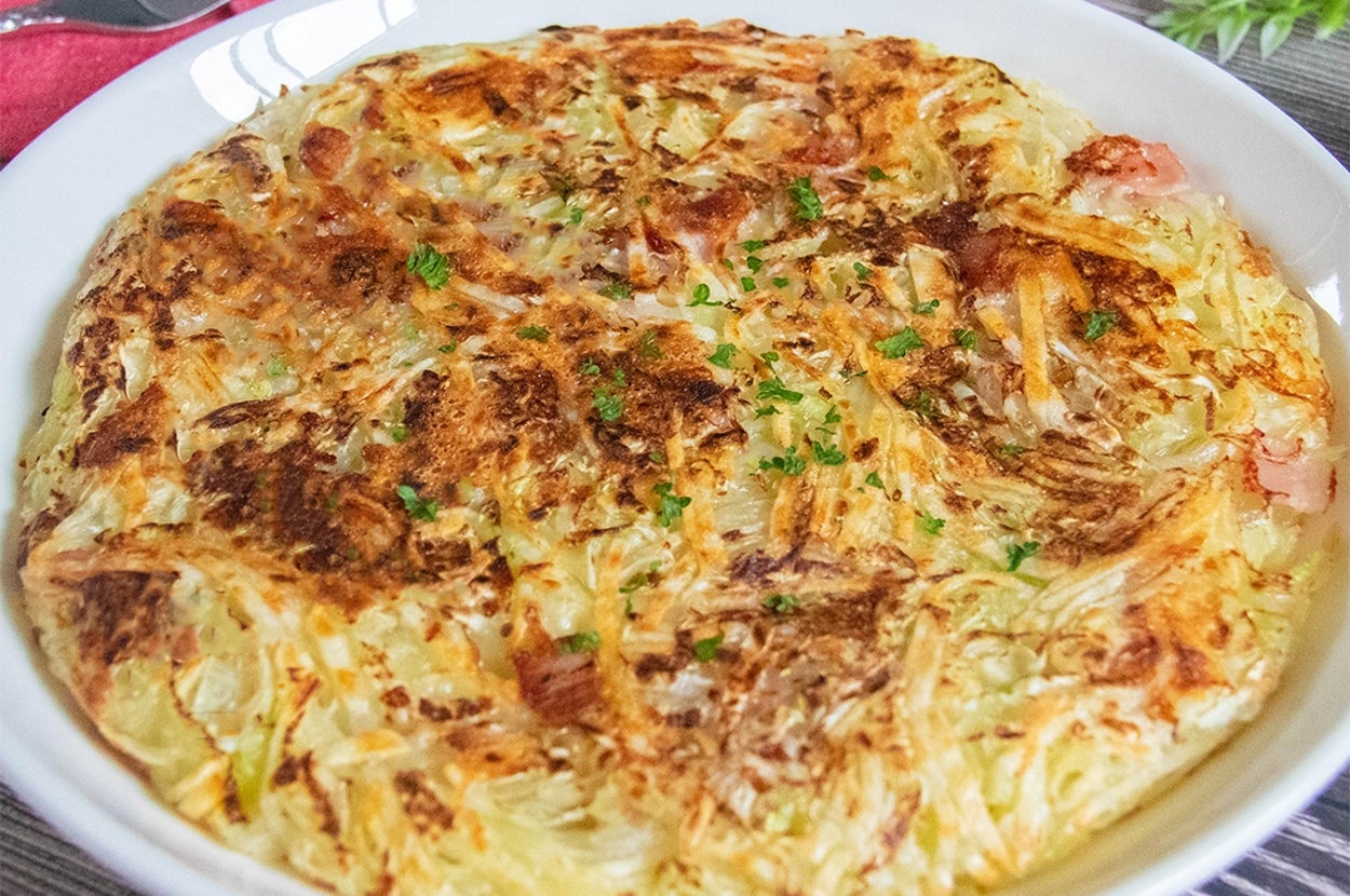 キャベツとじゃがいものガレットが白い皿に盛られている。チーズとベーコンも入っている。背景にはカトラリーと植物が見える。