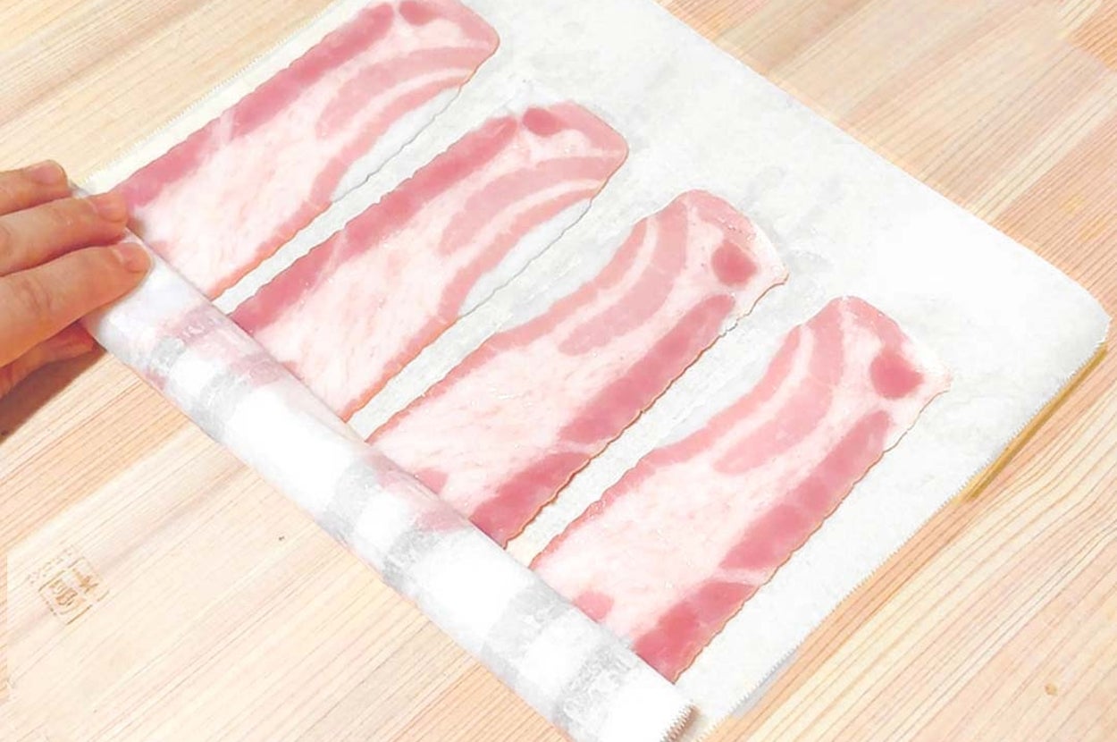 手が4枚のベーコンをキッチンペーパーで巻こうとしているところ。ベーコンは木のカウンターに置かれている。