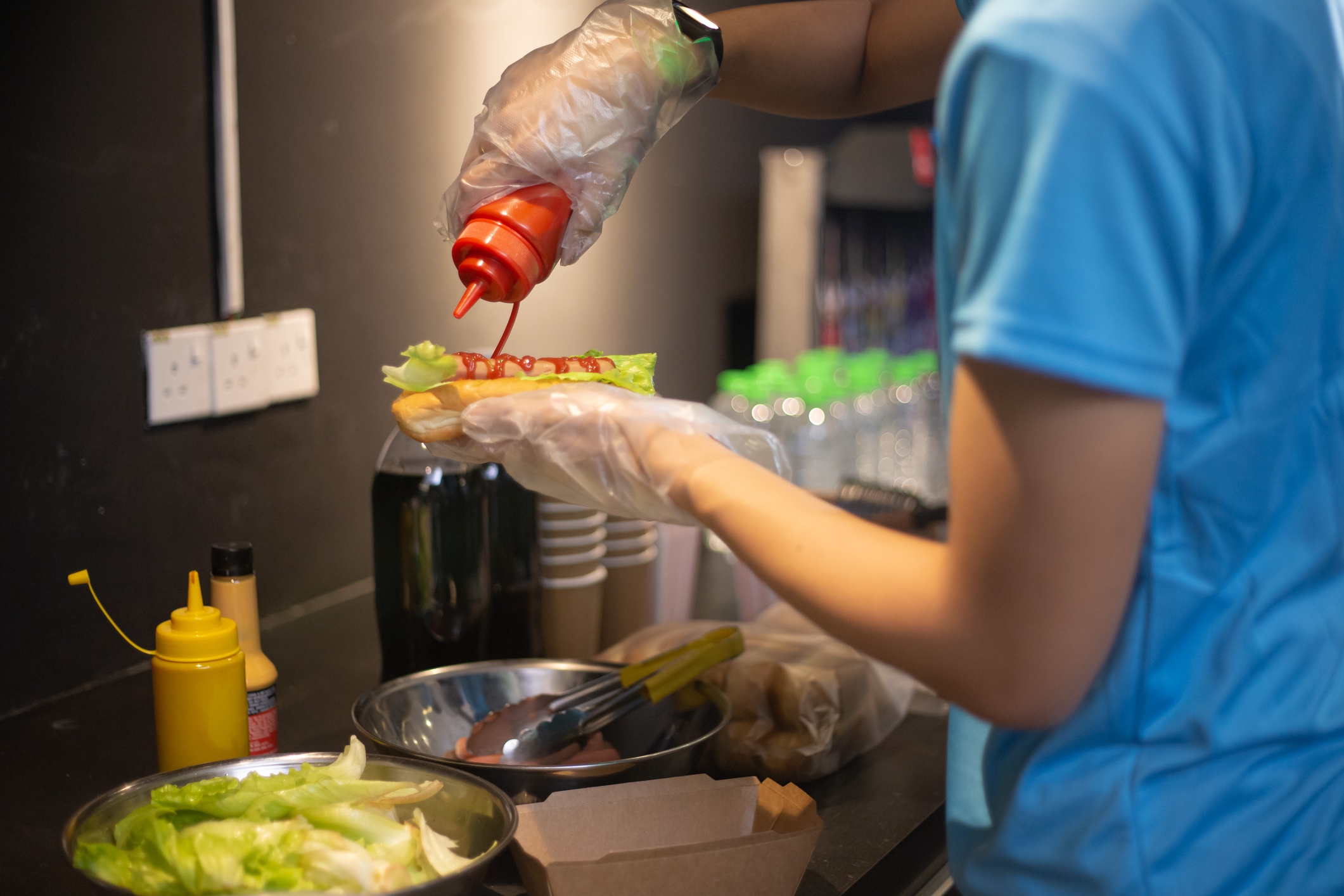 Person in a kitchen adds ketchup to a sandwich on a counter with condiments and bottled drinks. Wearing a blue shirt and plastic gloves