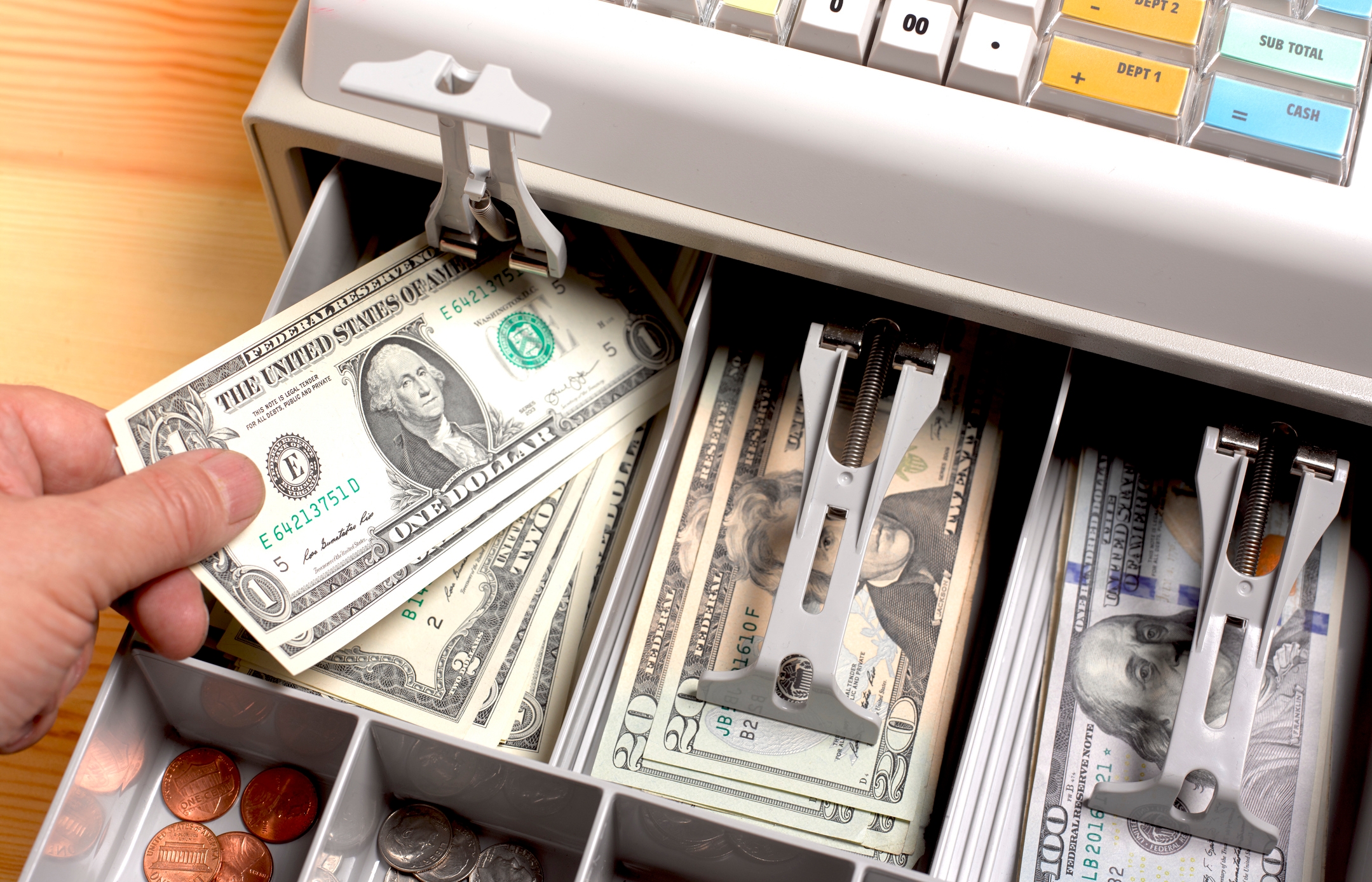 Hand placing a dollar bill in an open cash register drawer filled with various denominations of paper bills and coins