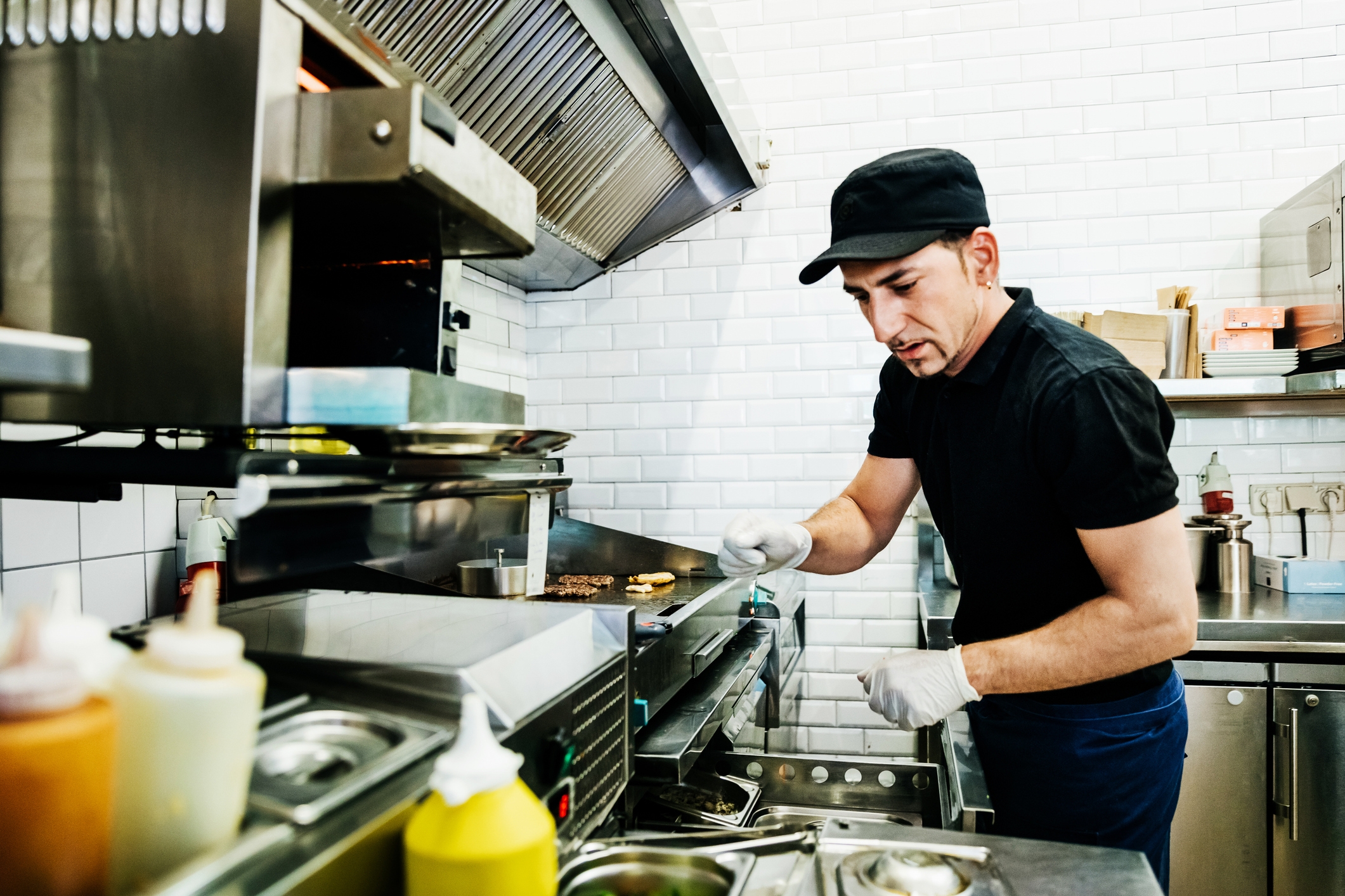 A chef wearing a black uniform and cap is focused on cooking at a grill in a professional kitchen