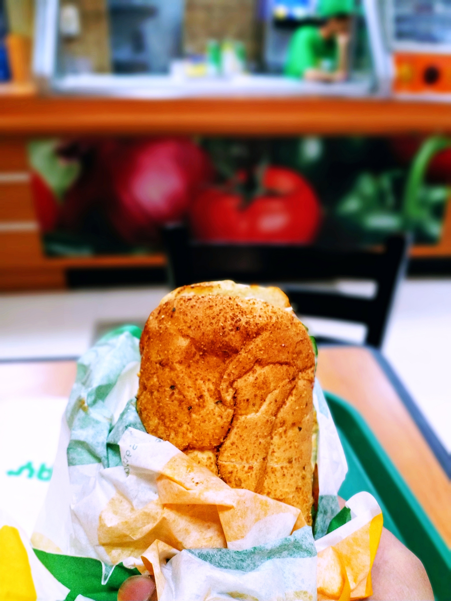 A sandwich wrapped in paper on a tray at a fast-food restaurant, with a blurred background of a counter and vegetables