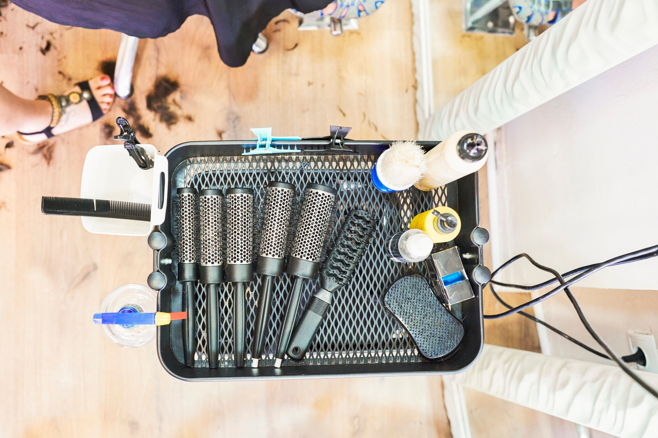 Salon cart with hairbrushes, spray bottles, and various hair styling tools, set on a wooden floor. Hair clippings are visible around the cart