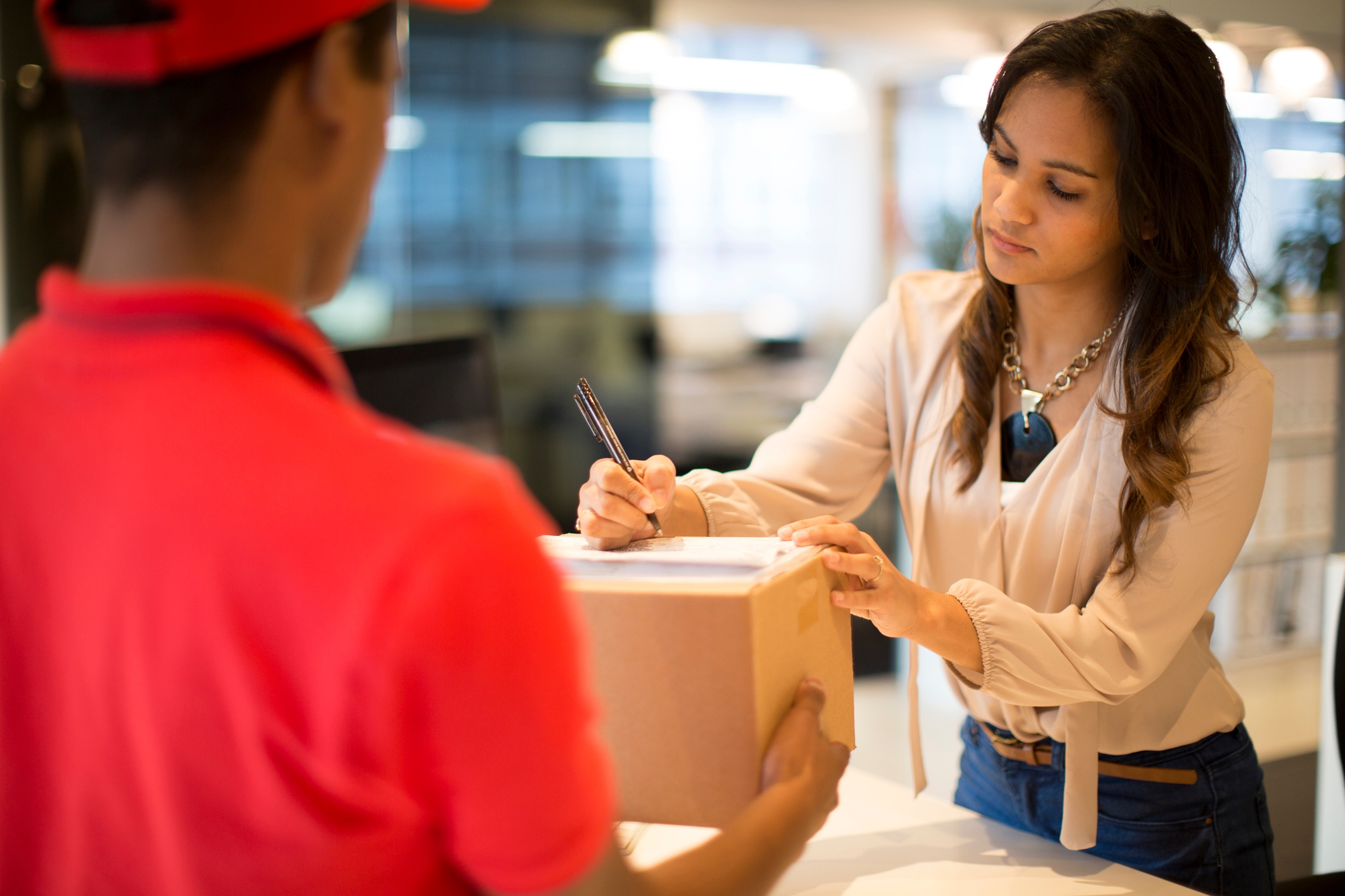 Woman signing a delivery package held by a courier inside an office, suggesting a workplace transaction or business delivery