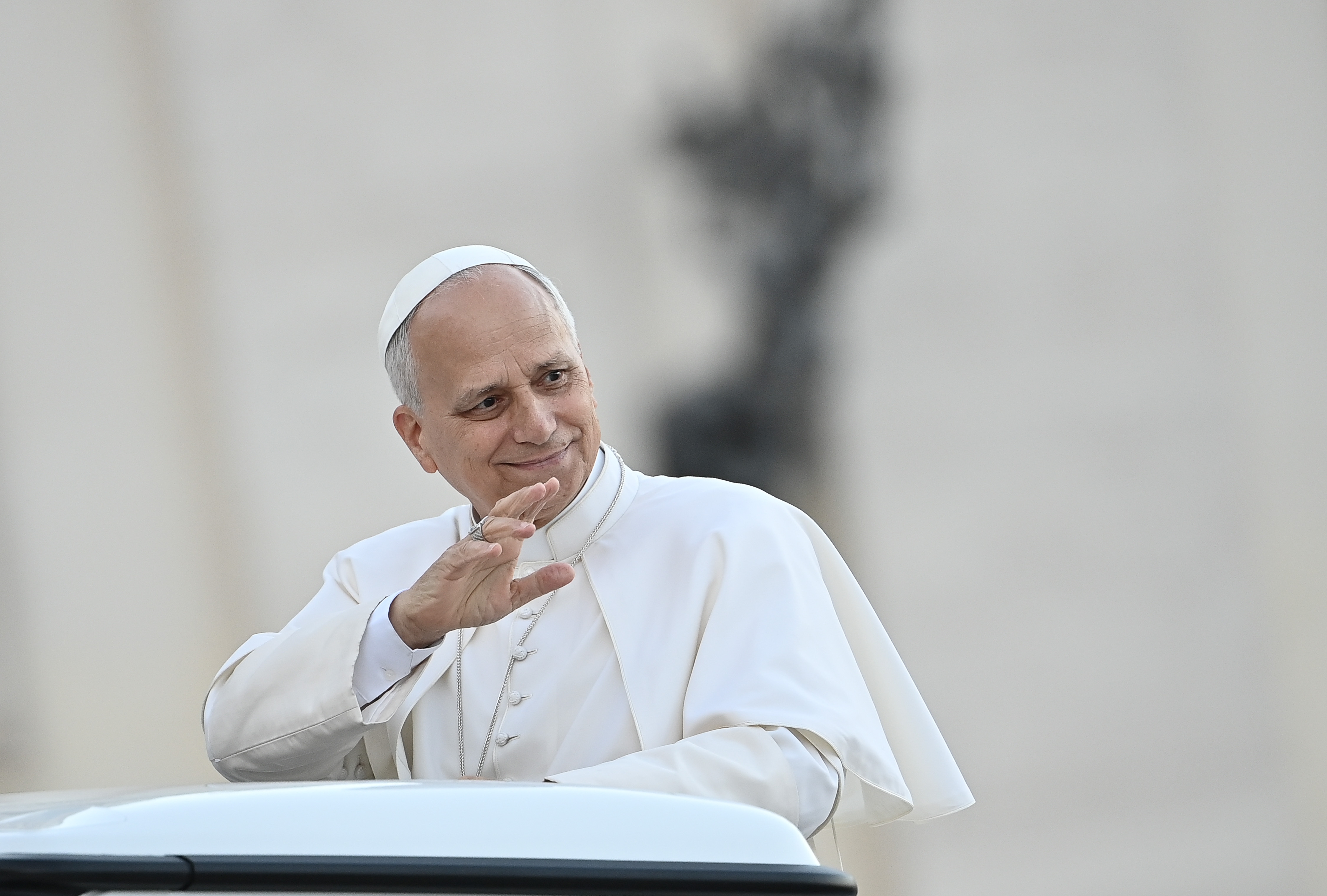 A man in a white robe and hat waves while standing in a vehicle
