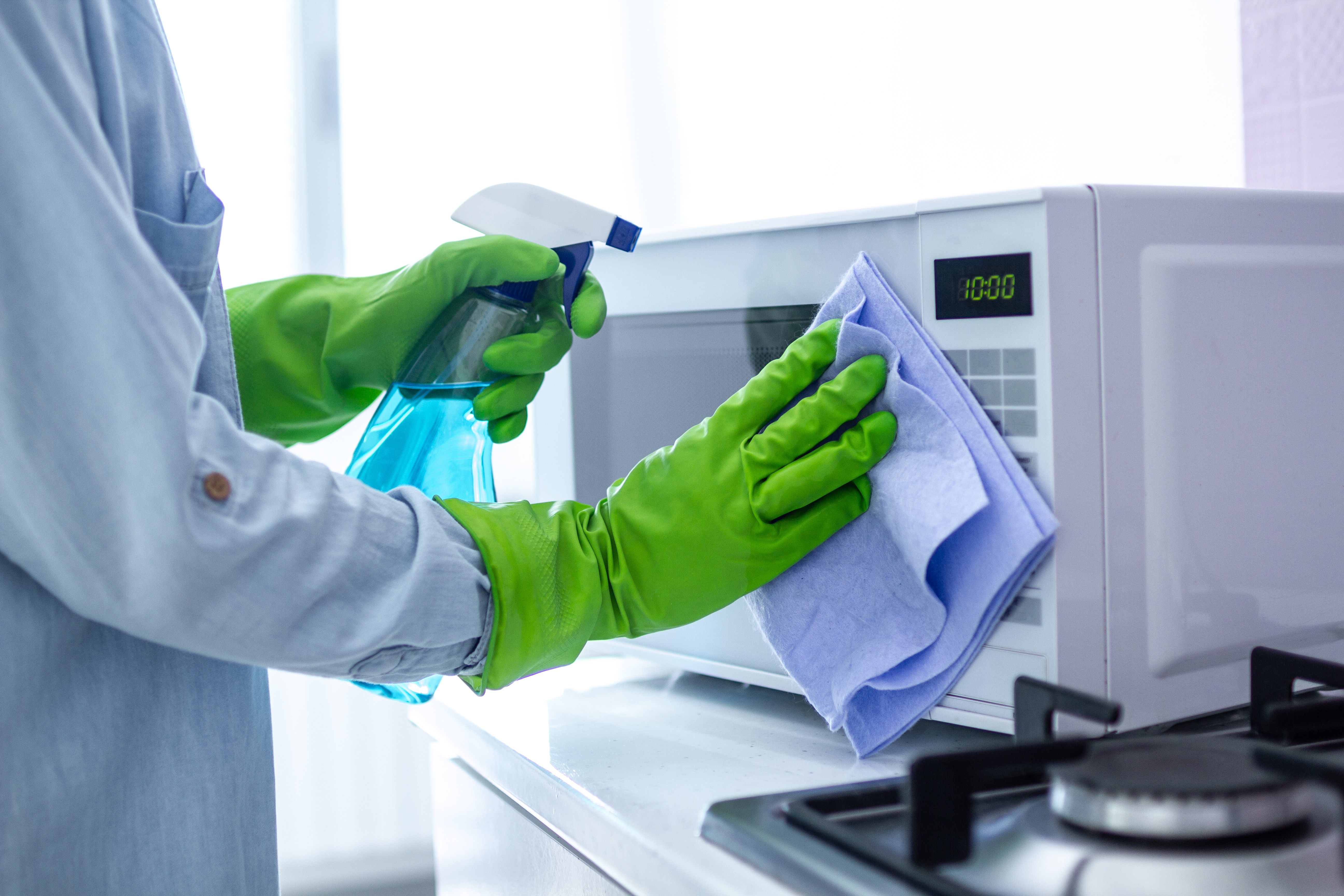 Person som rengjør mikrobølgeovn med spray og klut, iført grønne hansker. Gasskomfyr er i forgrunnen. Timer på mikrobølgeovn viser 10:00