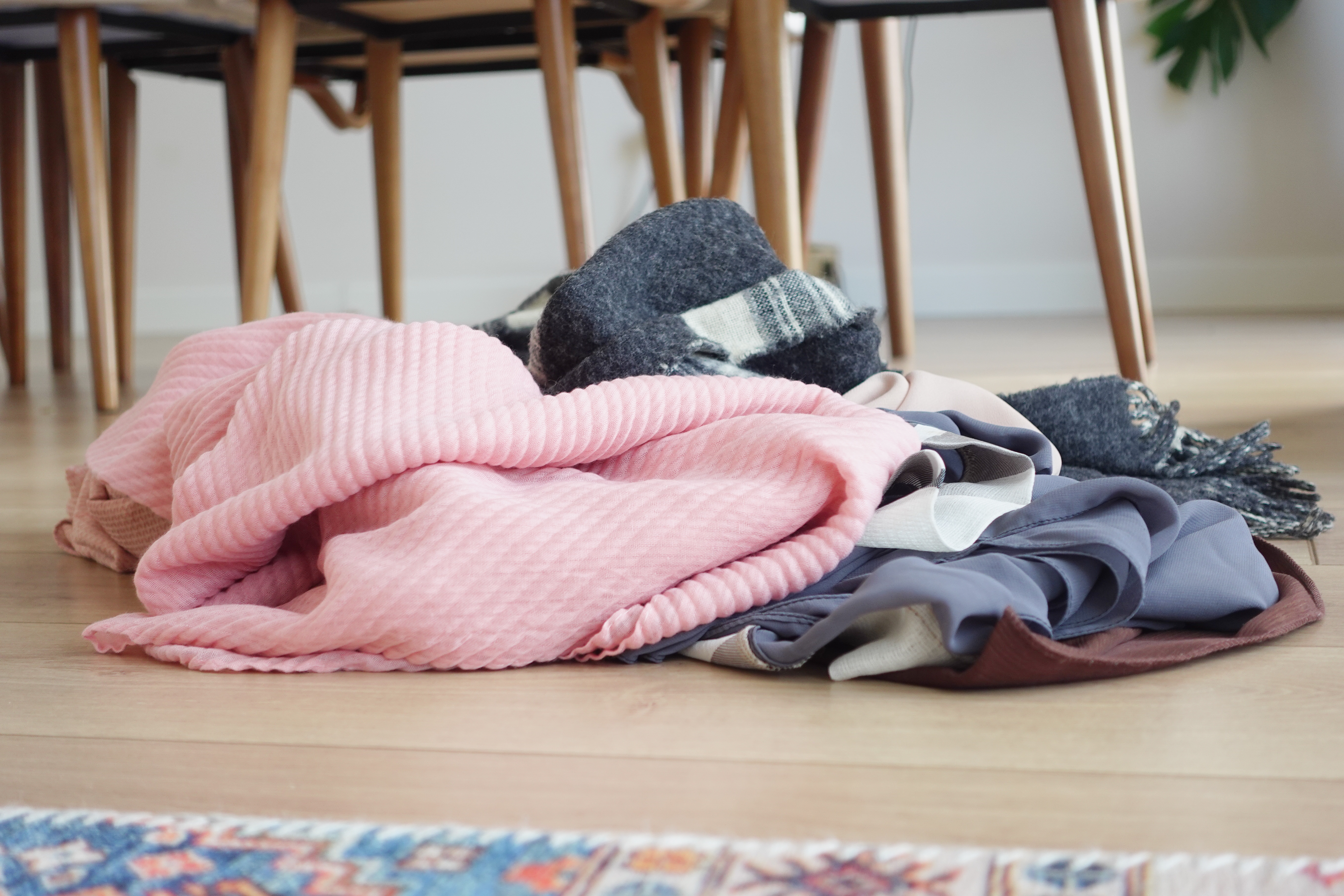 En haug med diverse klær og tepper på et tregulv, nær et spisebord med stoler