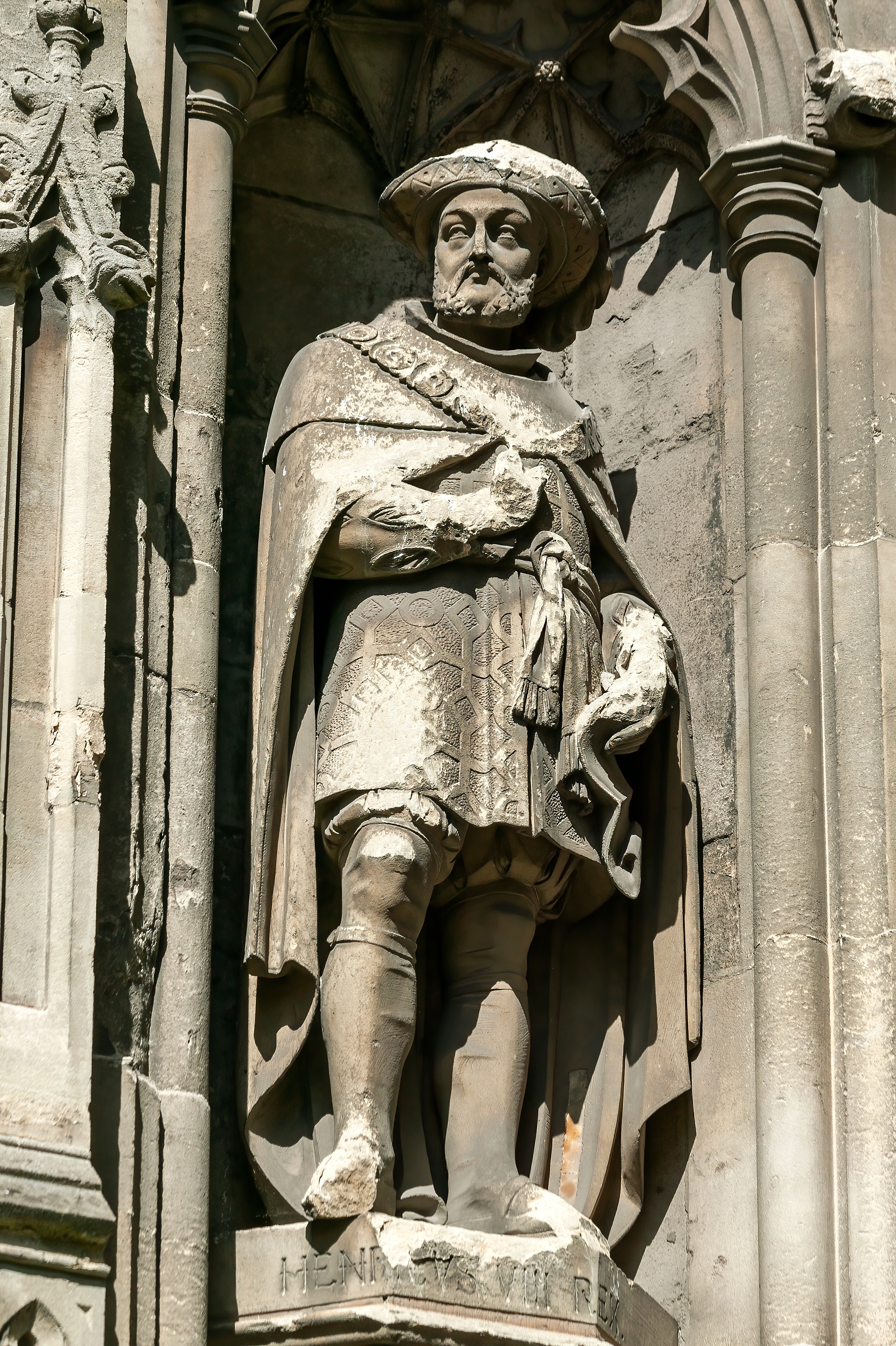 Stone statue of Henry VIII in traditional Tudor clothing, including a hat and cloak, set within a decorative architectural niche