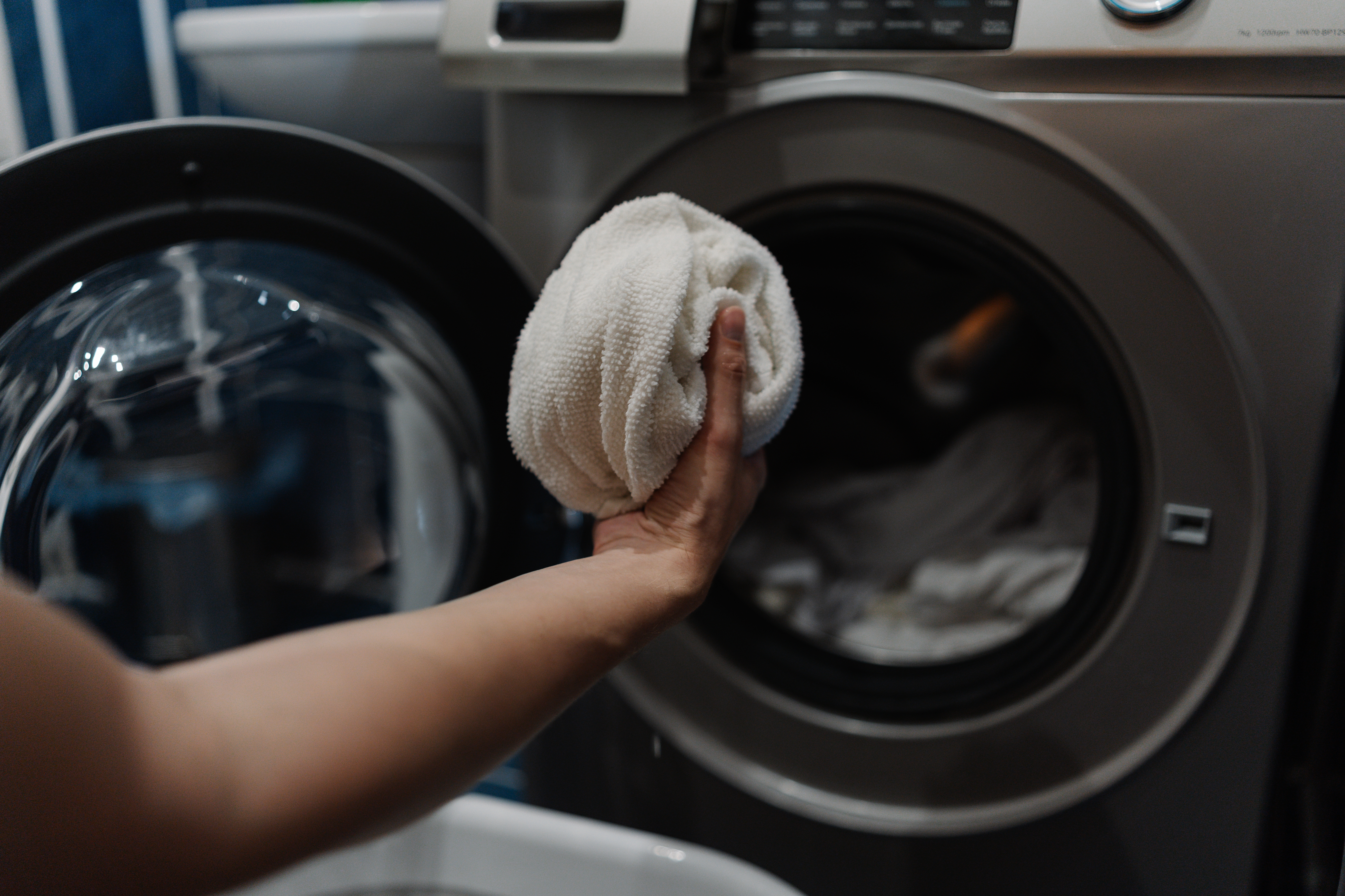 En person legger et hvitt håndkle inn i en åpen vaskemaskin i et vaskerom