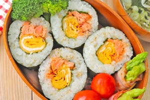 お弁当箱に寿司ロール、ブロッコリー、トマト、アスパラガスのベーコン巻きと味噌汁が盛り付けられている。