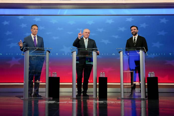 Three individuals in business suits stand at podiums on a stage set for a debate. The backdrop features a star pattern
