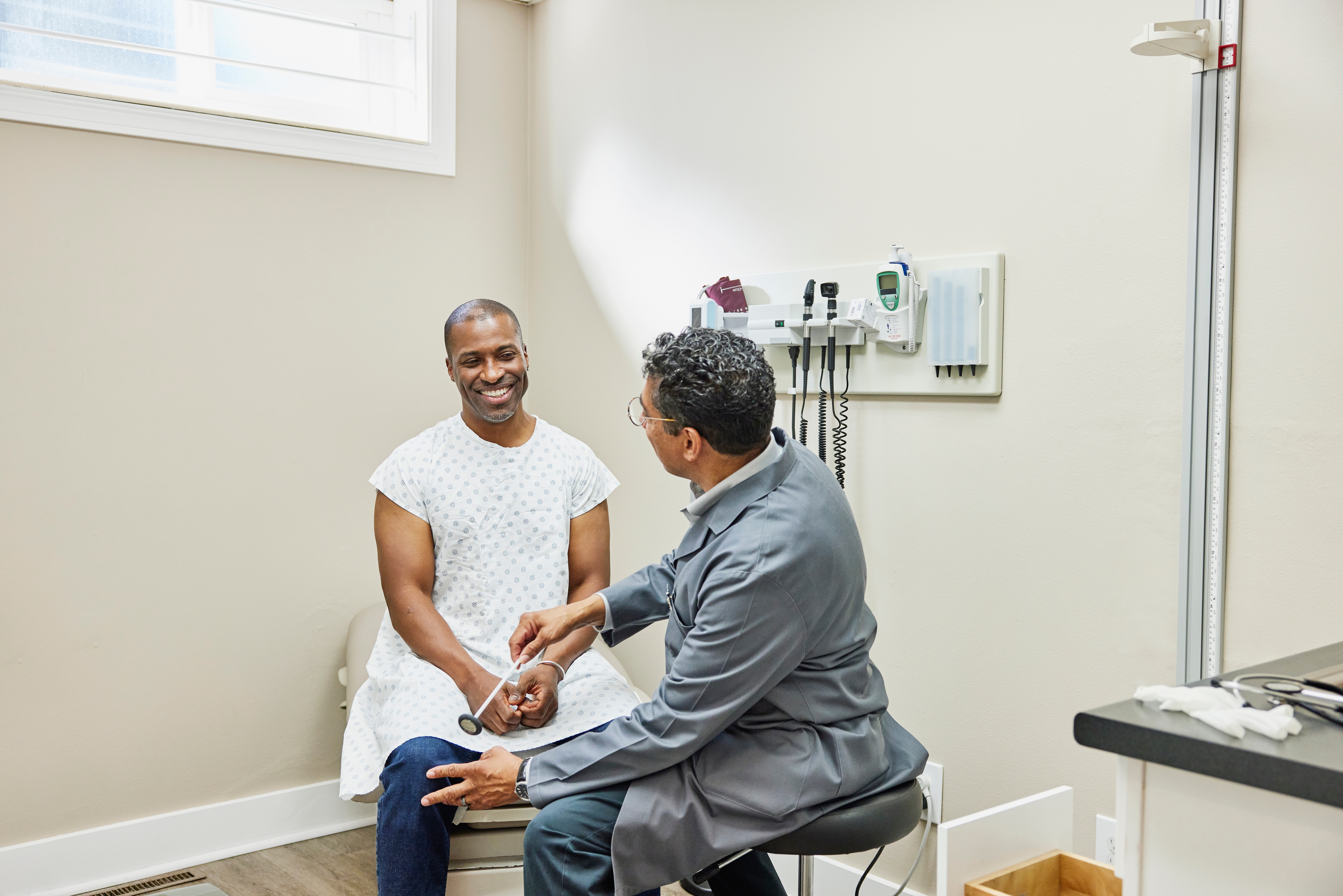 En lege i hvit frakk snakker med en smilende pasient i en sykehuskjole under en konsultasjon på et legekontor