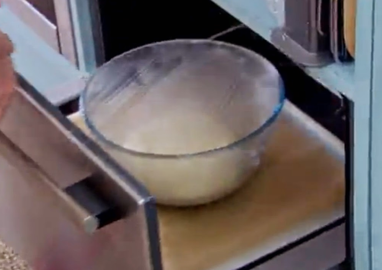 A bowl covered with plastic wrap is placed inside a warming drawer, showing a dough proofing process