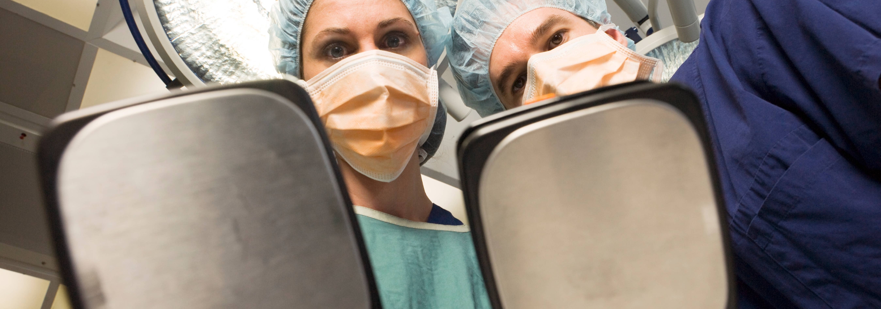 Two medical professionals in scrubs and masks hold defibrillator paddles, viewed from the patient's perspective in an operating room