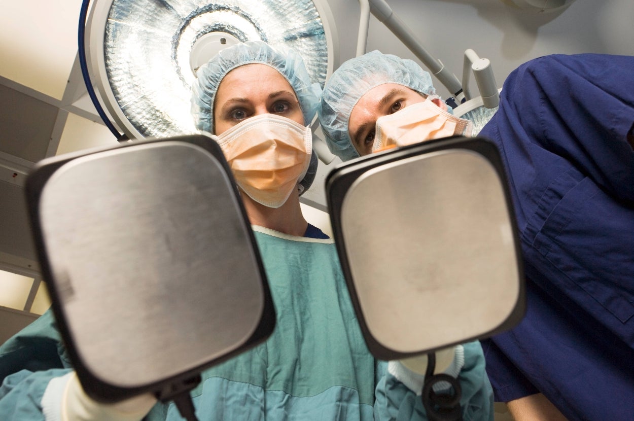 Two medical professionals in scrubs and masks hold defibrillator paddles, viewed from the patient's perspective in an operating room
