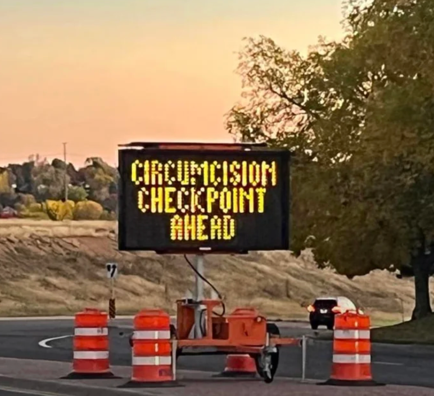Humorous road sign reads "Circumcision Checkpoint Ahead" near a curved road with traffic cones