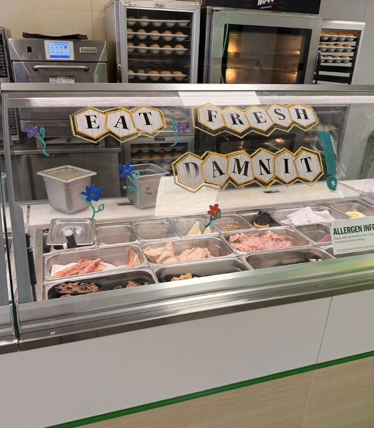Sandwich shop counter with a humorous sign that says "Eat Fresh Damnit" above trays of various deli meats and toppings