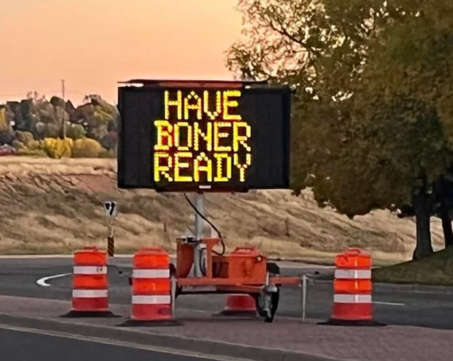 A road sign displays humorous text, "Have Boner Ready," surrounded by orange traffic barrels on a roadside