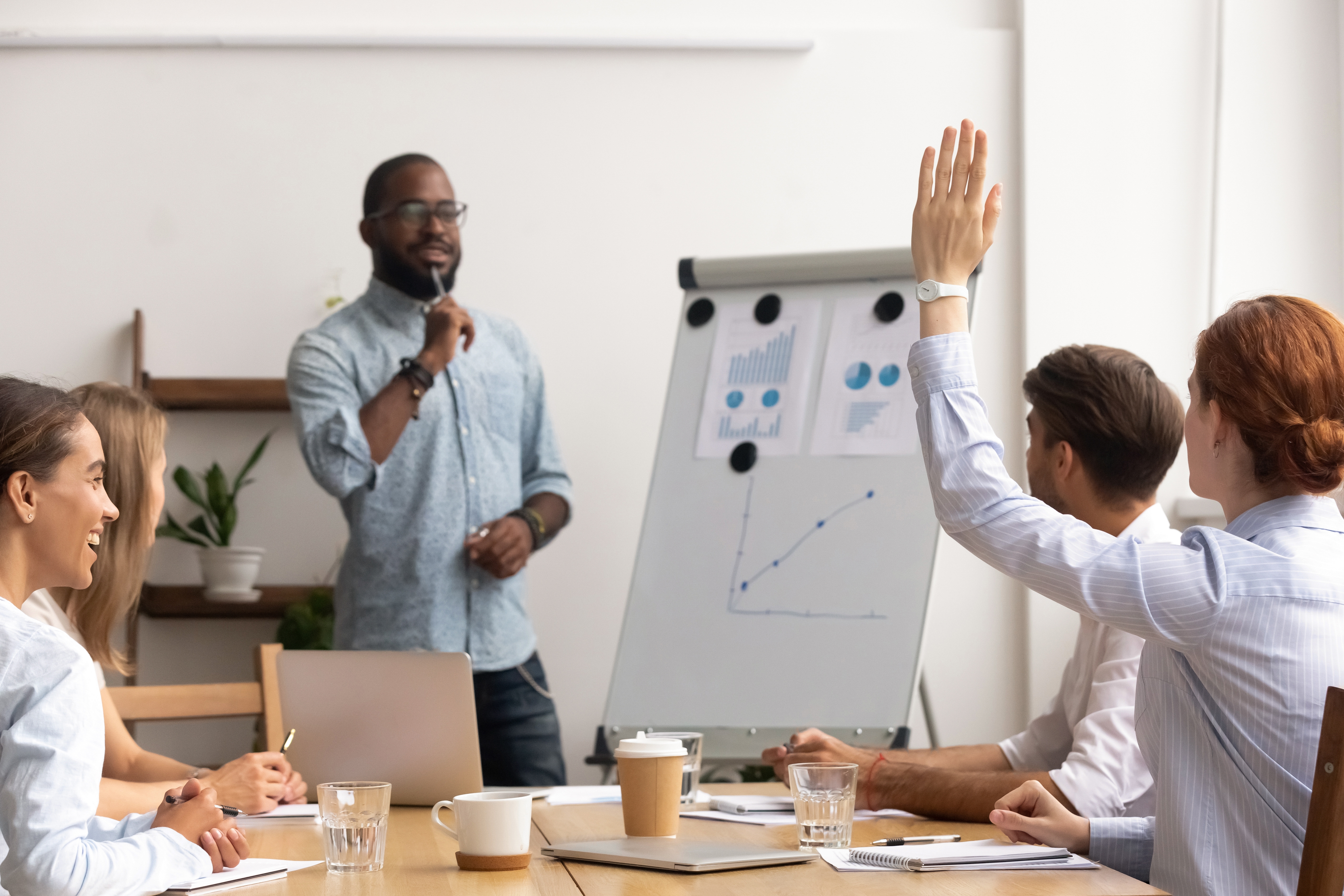 En person presenterer for en liten gruppe i et møterom. En løftet hånd indikerer deltakelse. Diagrammer og notater er synlige på en flippover