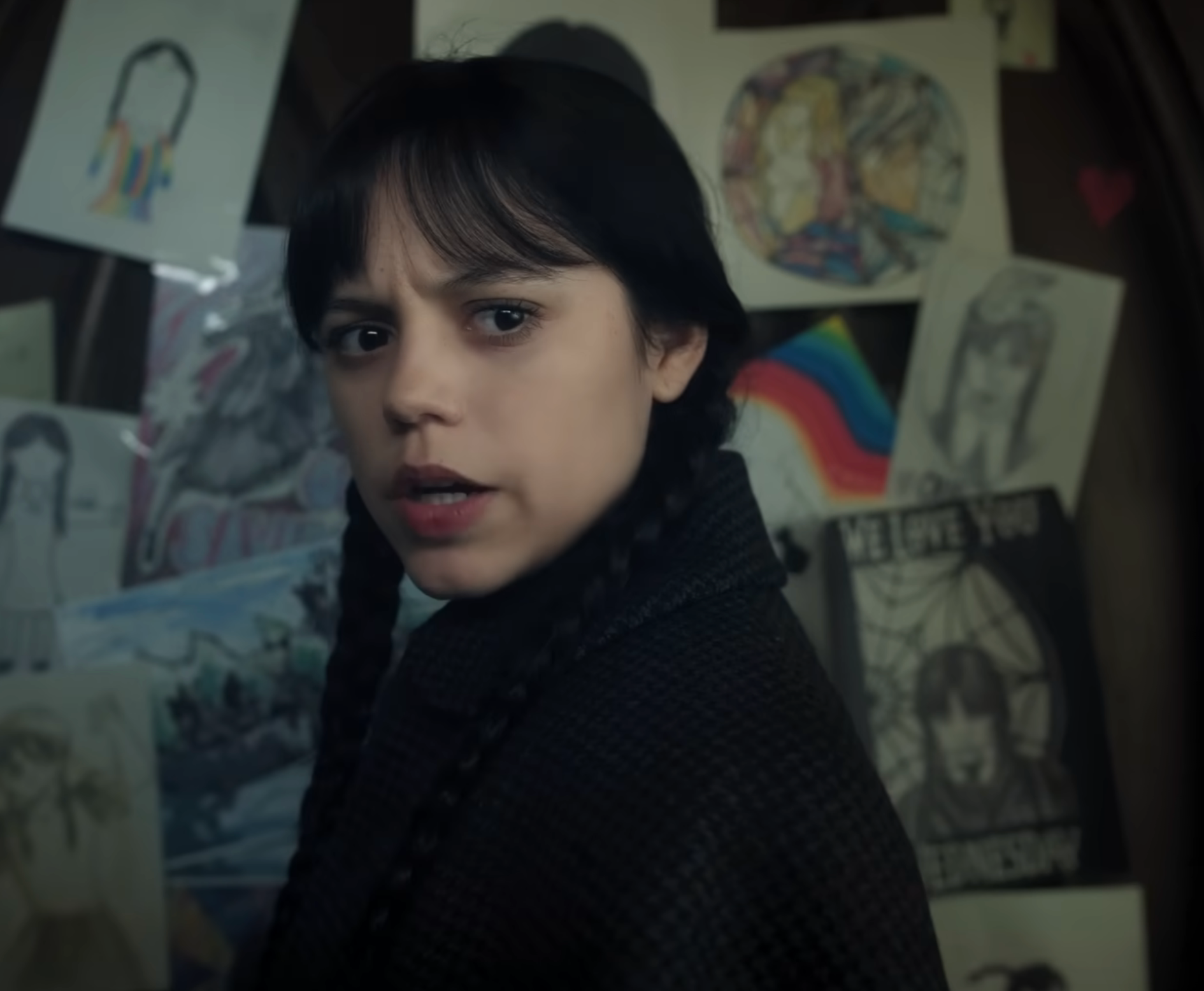 Wednesday with long braided hair stands in front of a wall filled with various drawings and art